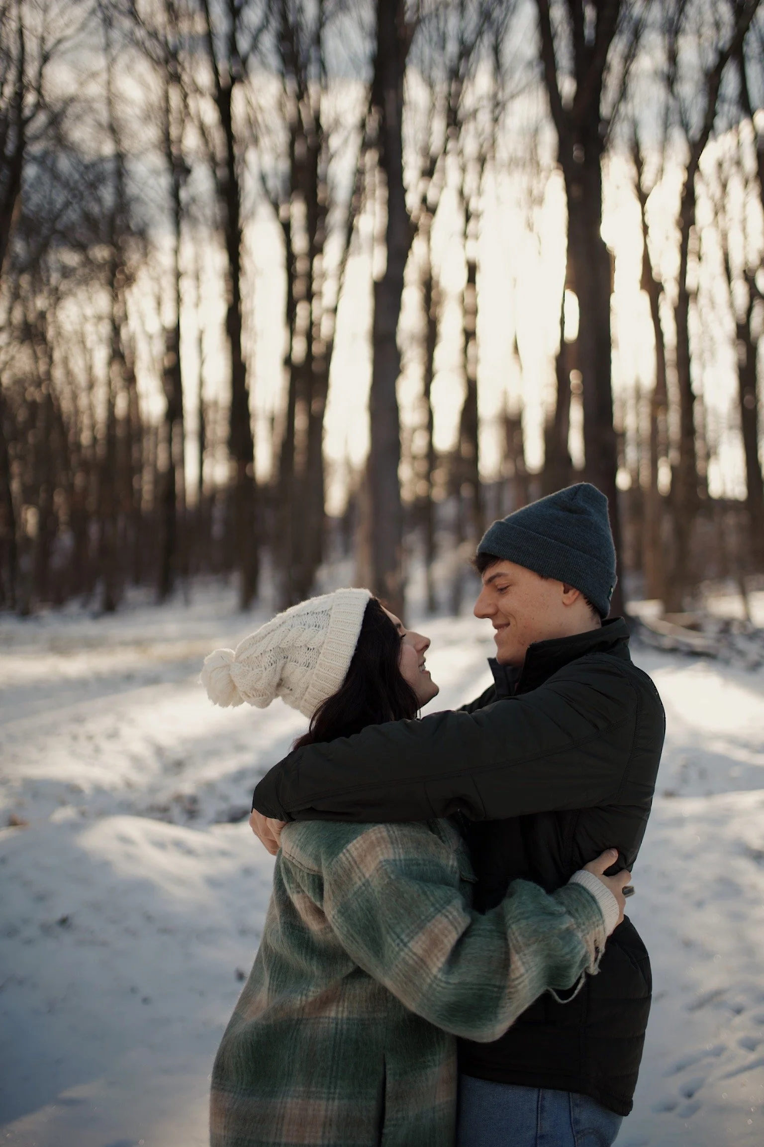 A couple hugging outdoors in a snowy forest during winter, wearing warm clothing and hats.