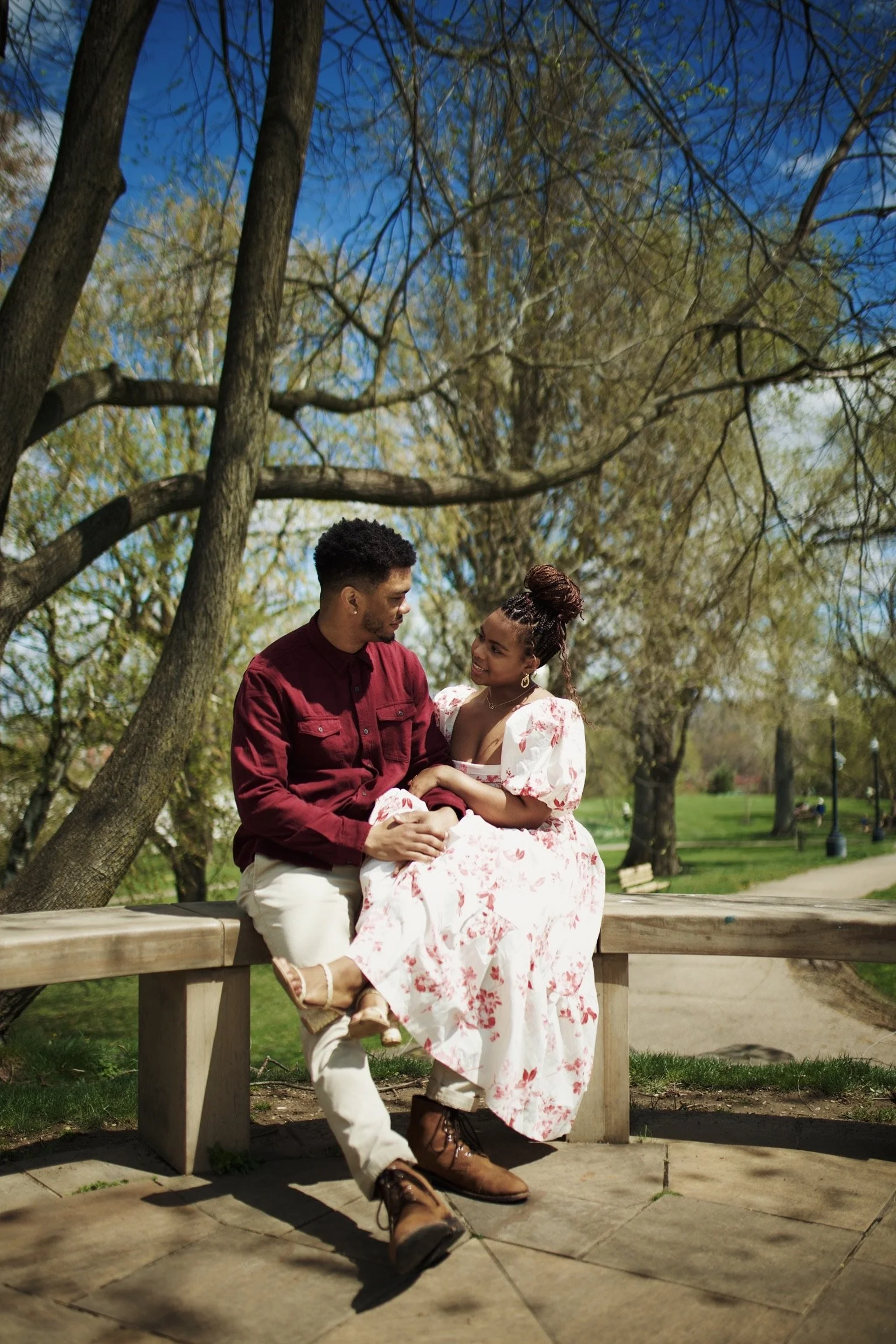 A couple sitting on a park bench under a tree, smiling and looking at each other, with a park background on a bright, sunny day.