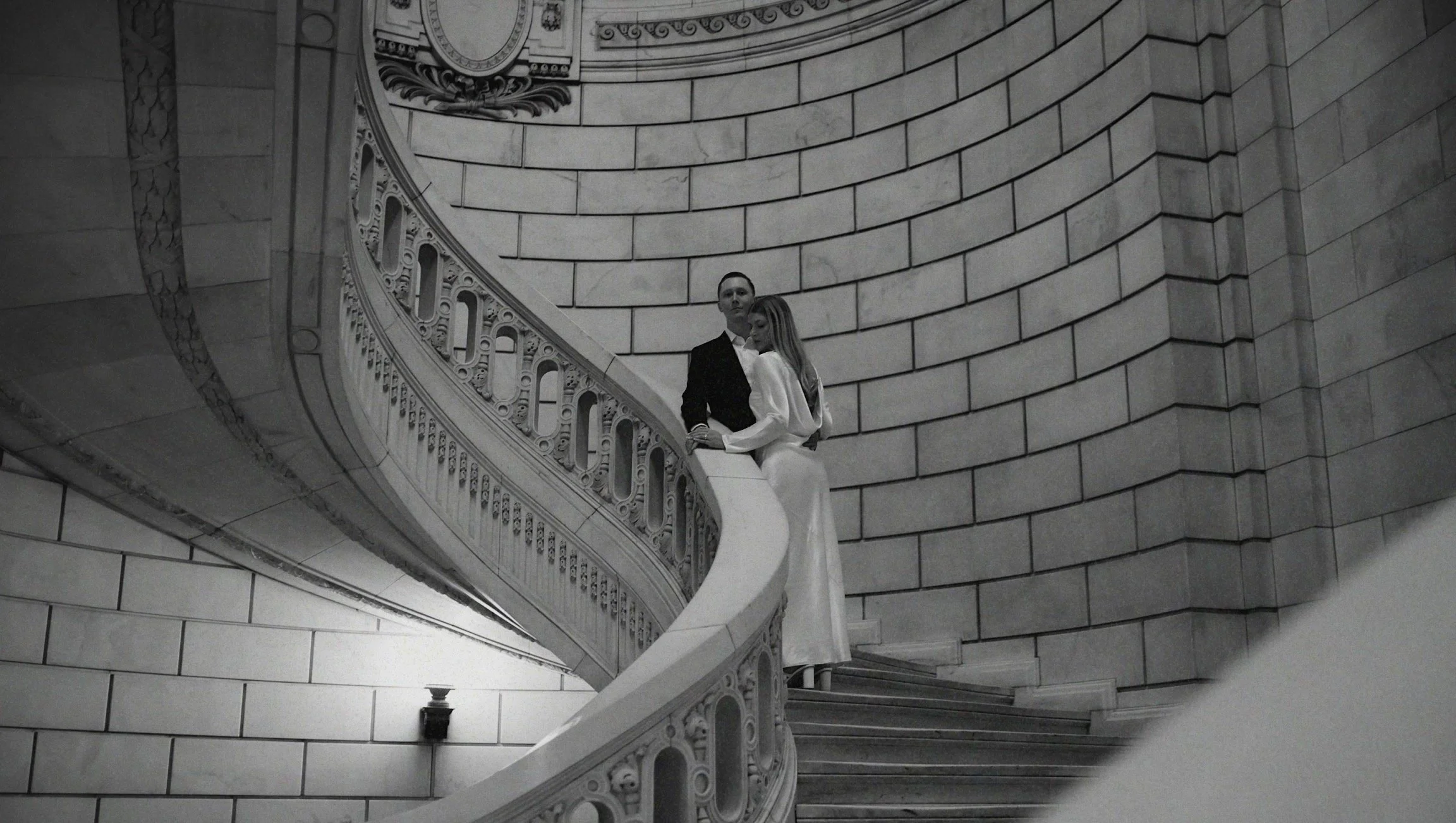 Black and white photo of a man in a suit and a woman in a flowing dress standing on a curved, ornate stone staircase in a grand building with a stone wall background.
