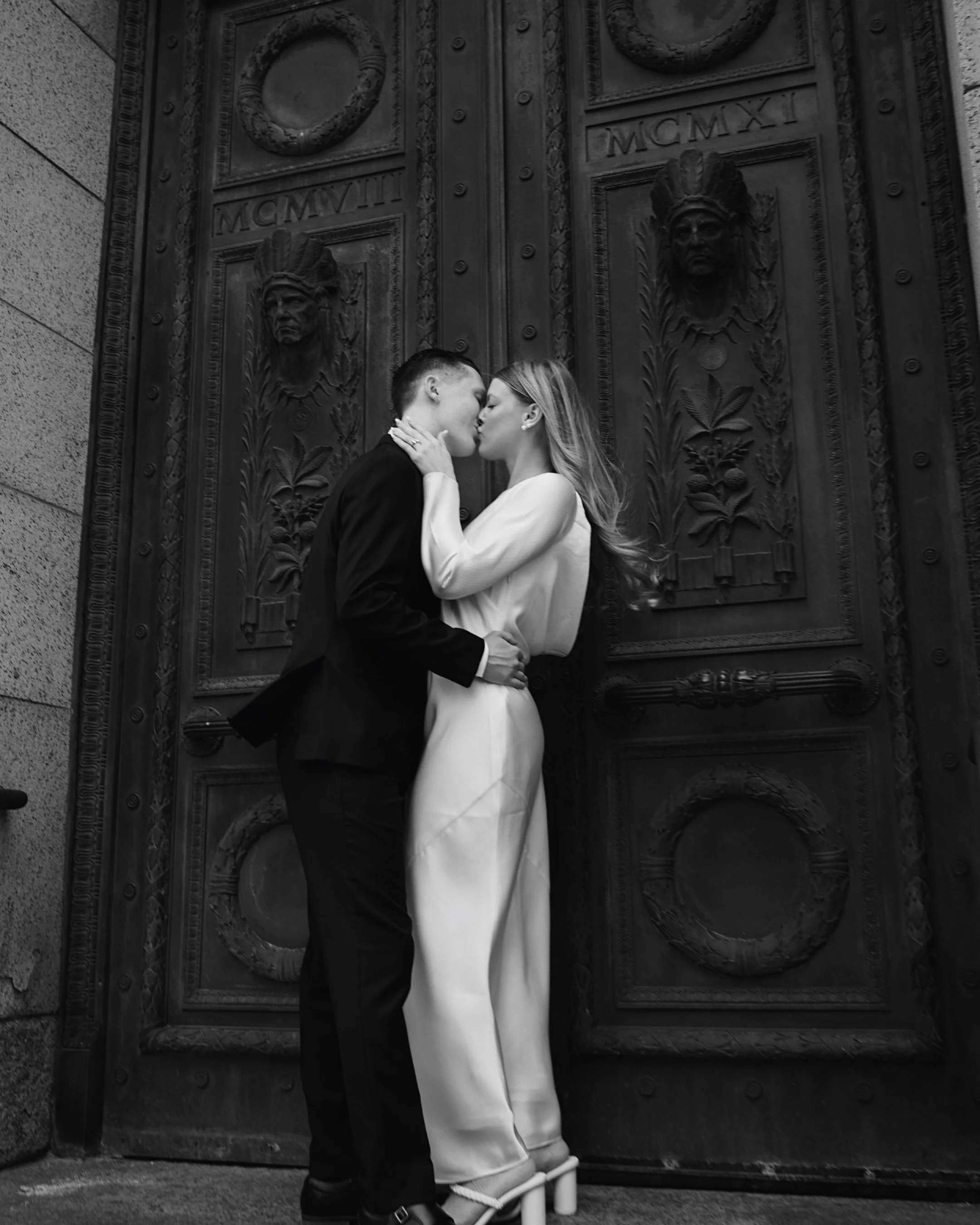 A black-and-white photo of a couple kissing in front of an ornate, large metal door with carved faces and decorative details.
