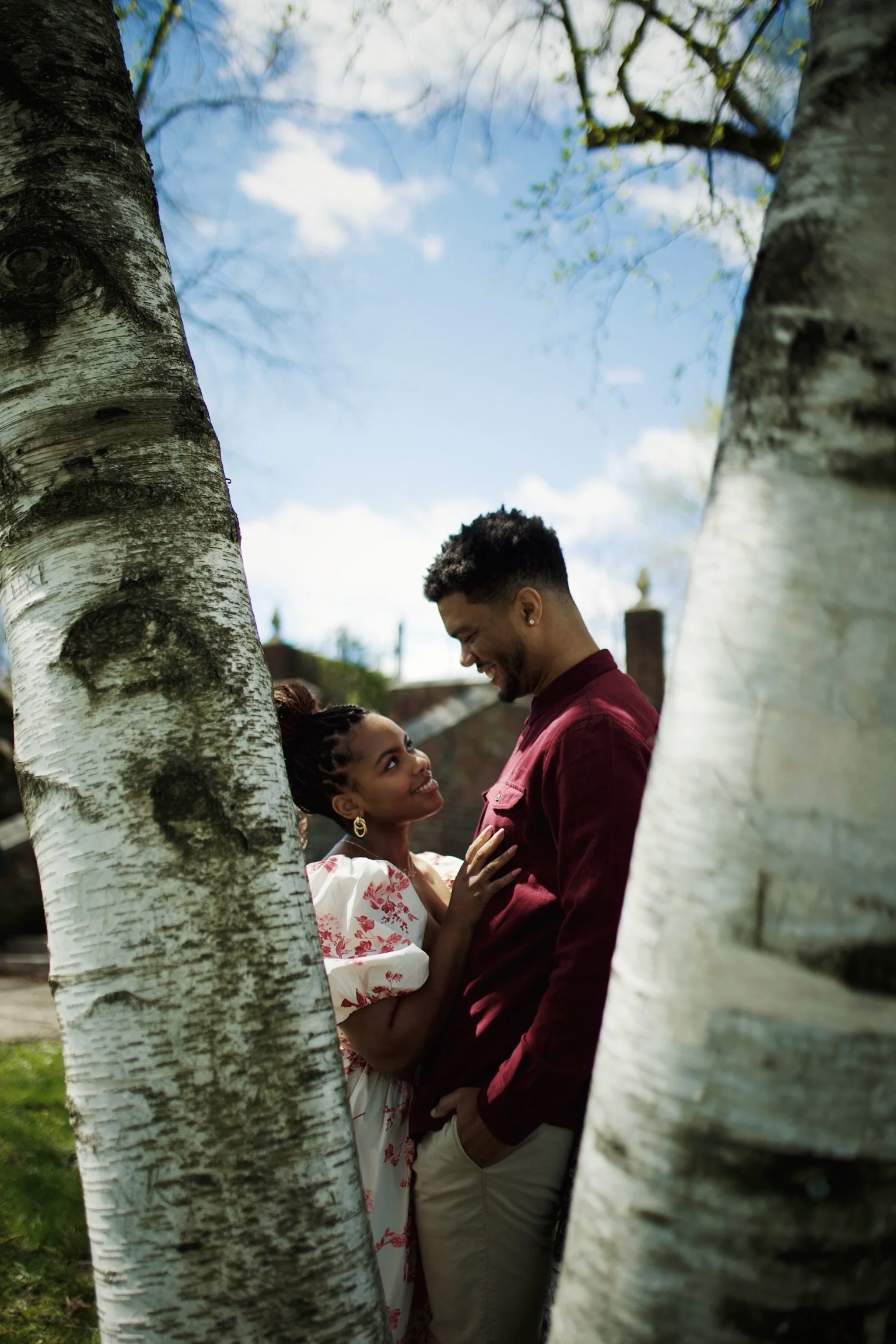 A man and woman standing close together, smiling and looking into each other's eyes, framed by two birch trees outdoors on a sunny day.