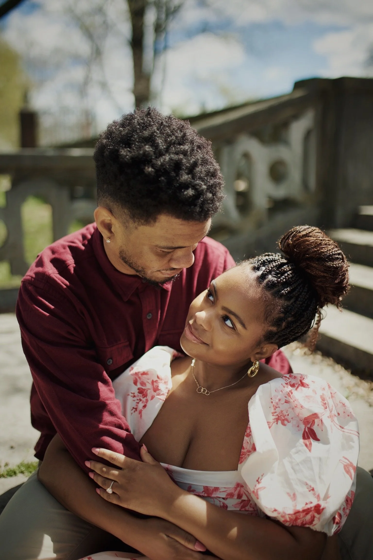 A couple sitting outdoor on a wooden surface, gazing into each other's eyes with their faces close. The woman is wearing a white and red floral dress with puffed sleeves, gold earrings, a gold necklace, and has a hair bun. The man is wearing a burgun