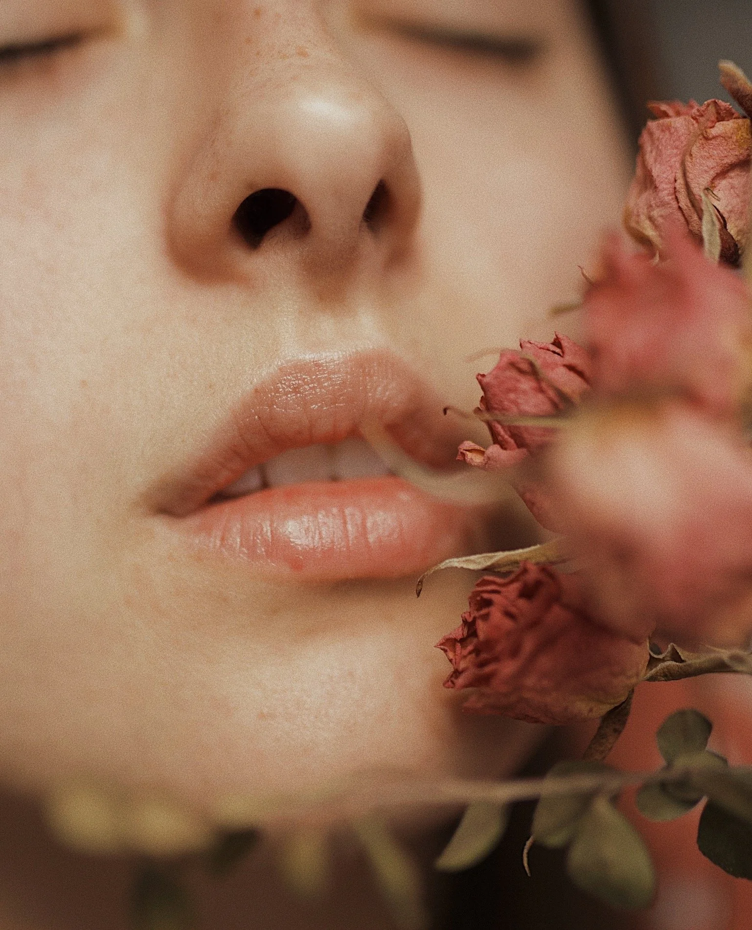 Close-up of a woman's face, focusing on her nose and lips, with dried pink flowers near her mouth.