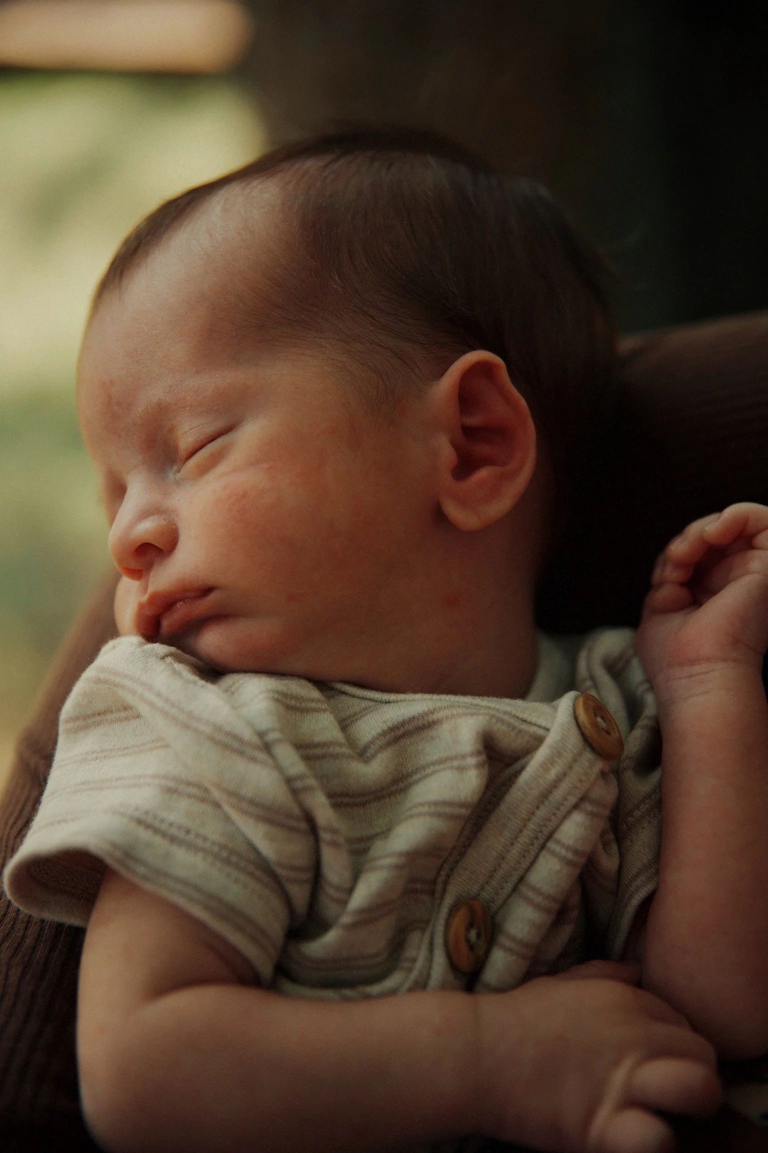 A sleeping baby with short dark hair, wearing a beige and brown striped shirt with buttons, resting against someone's arm.