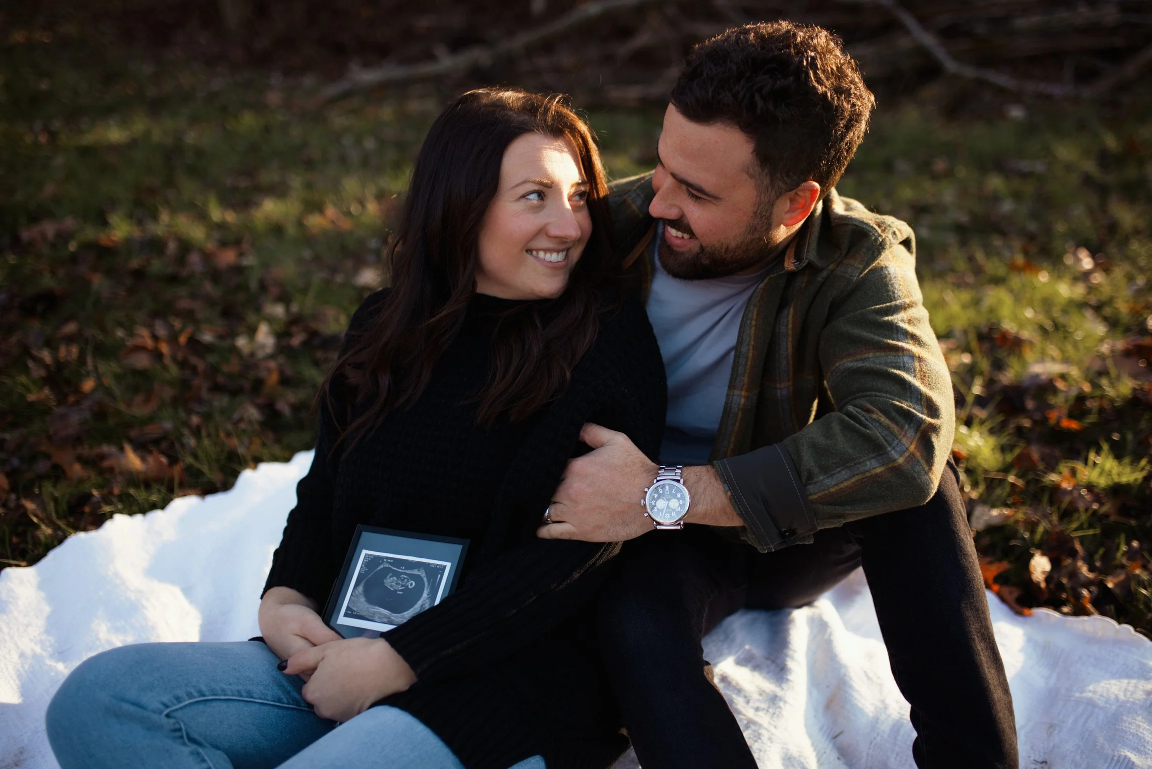 A couple sitting on a white blanket outdoors, smiling at each other. The woman is holding an ultrasound picture. The man is wearing a watch and plaid shirt, and the woman has dark hair and a black sweater.