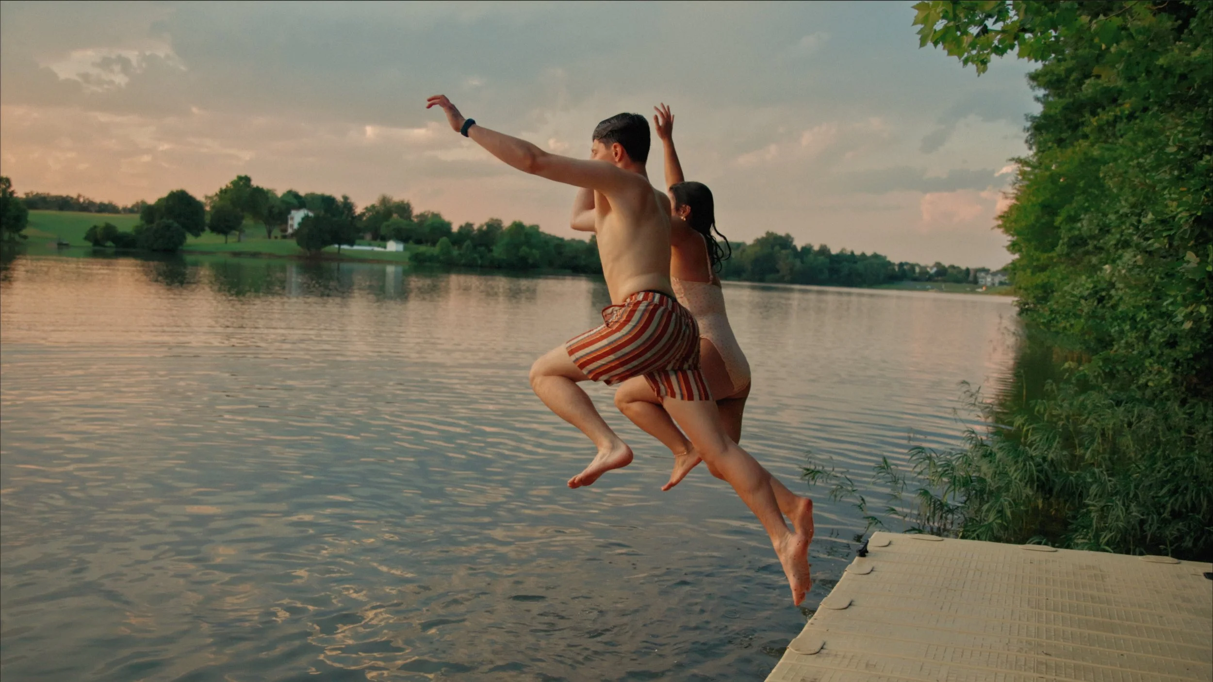 A man and woman jumping off a dock into a lake at sunset, surrounded by trees and a green landscape.