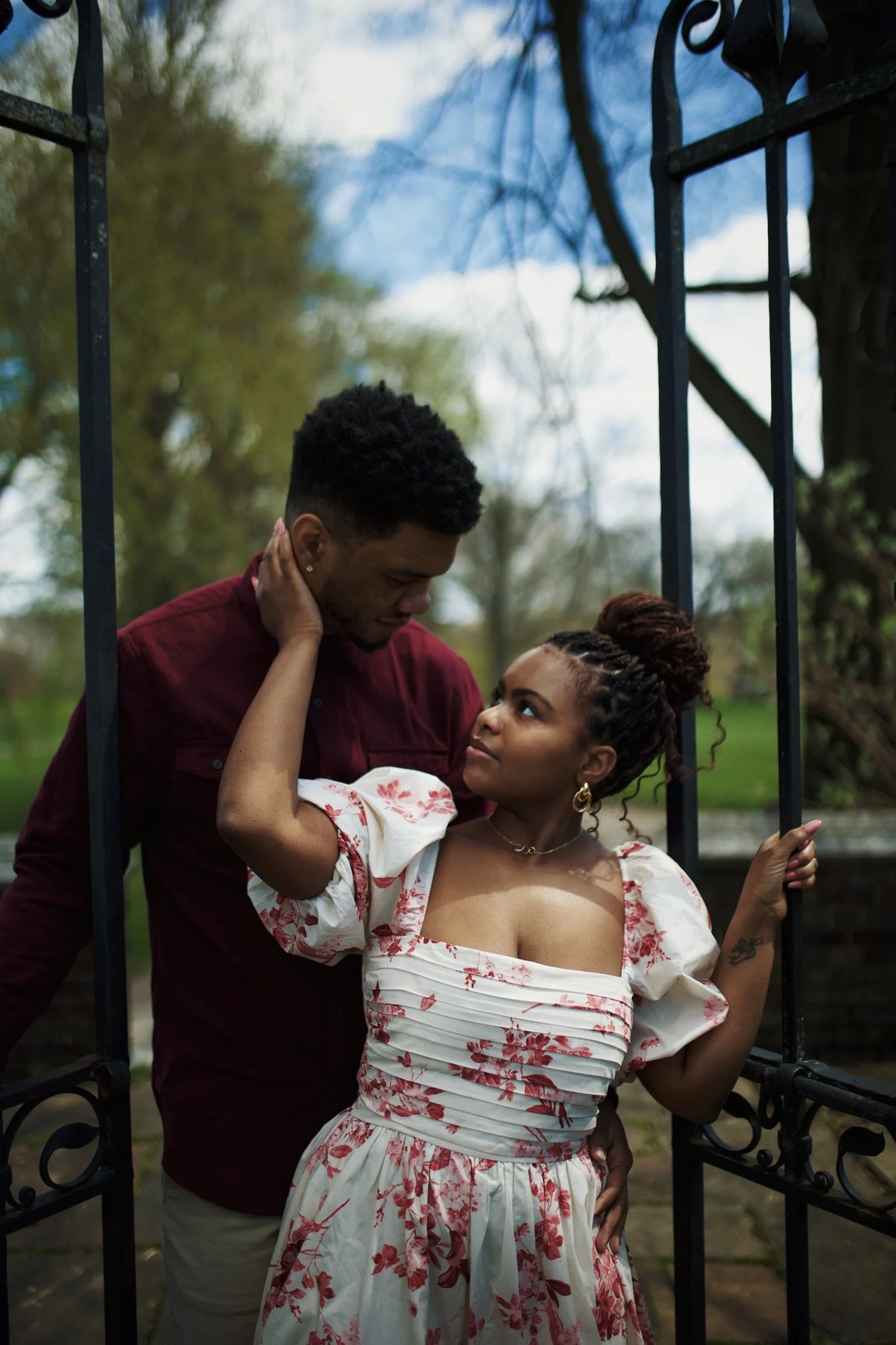 A man and woman stand close together outdoors, gazing into each other's eyes. The woman, with curls in a bun, is wearing a white dress with pink floral prints and gold jewelry. The man, with short curly hair, is wearing a dark red shirt. The scene is