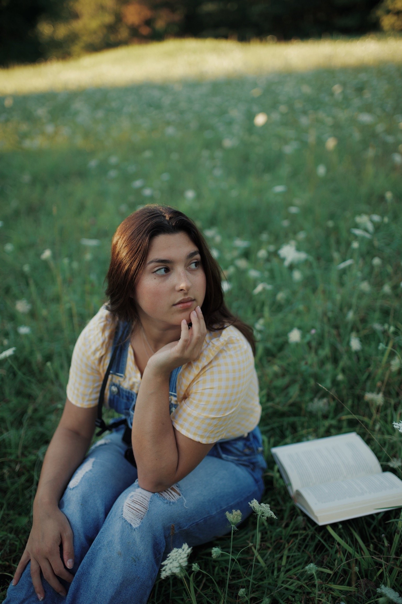 A young woman with brown hair, wearing a yellow checkered shirt and ripped jeans, sitting in a field of grass and white flowers, looking to the side with her hand on her chin, with an open book beside her.