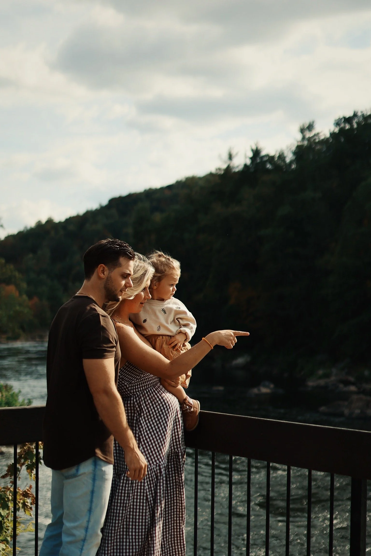 A family of three standing on a bridge overlooking a river, with a forested hillside in the background. The woman is holding a young girl, and the girl is pointing toward the distance.