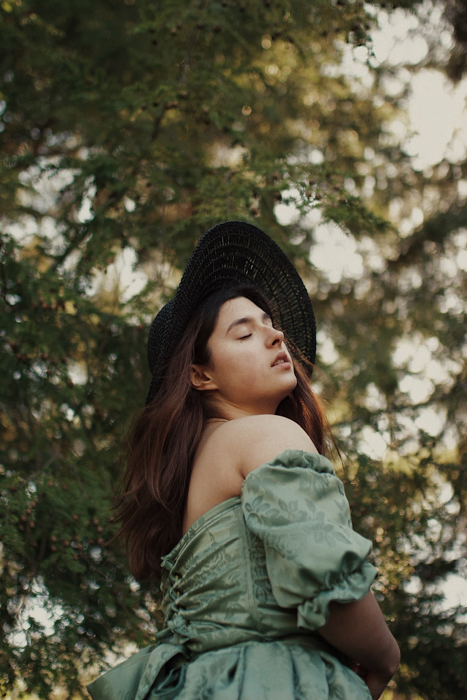 A woman with long brown hair wearing a black wide-brimmed hat and a green off-the-shoulder dress with puffed sleeves standing outdoors among trees, with her eyes closed and head tilted back.
