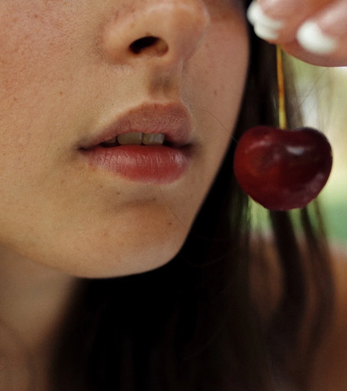 Close-up of a woman's face near her lips and nose, with a cherry hanging close to her face.