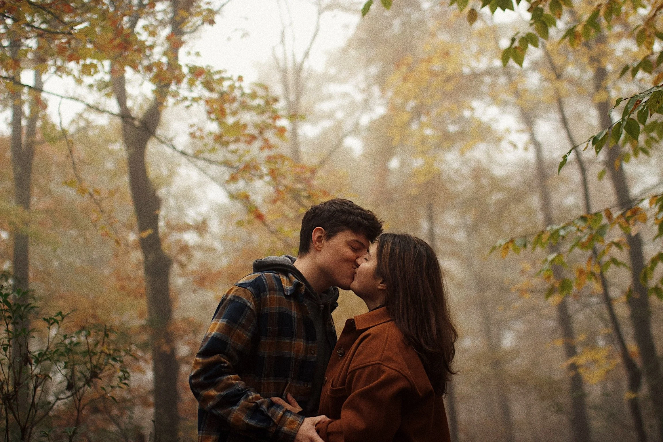 A couple kissing in a foggy autumn forest surrounded by trees with fall foliage.
