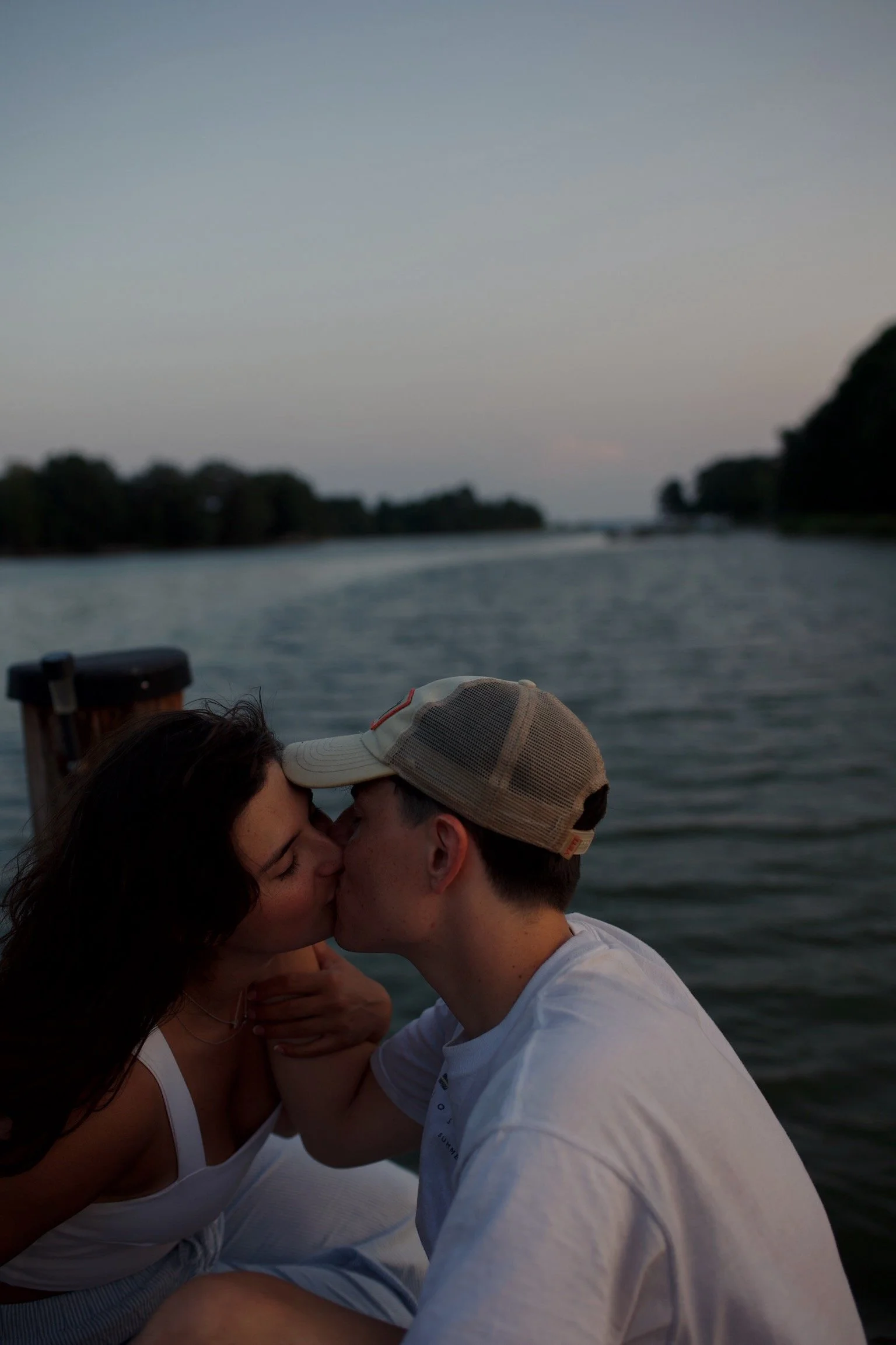 A couple sharing a kiss on a boat during sunset, with water and trees in the background.