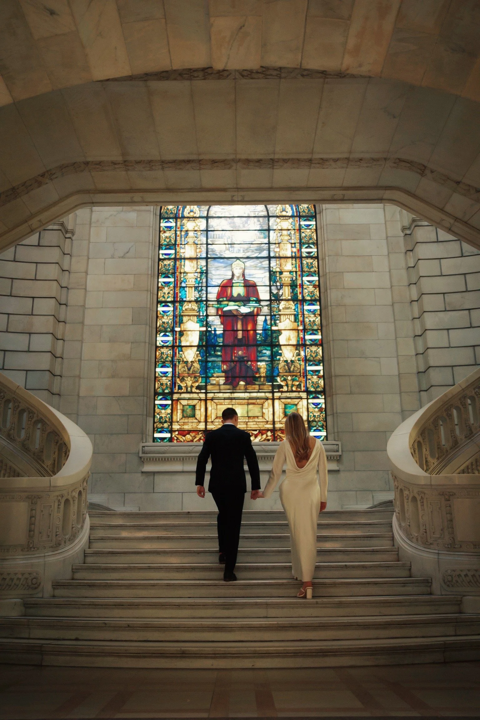 A man and woman walking up a marble staircase holding hands inside a building with a large stained glass window of a religious figure behind them.