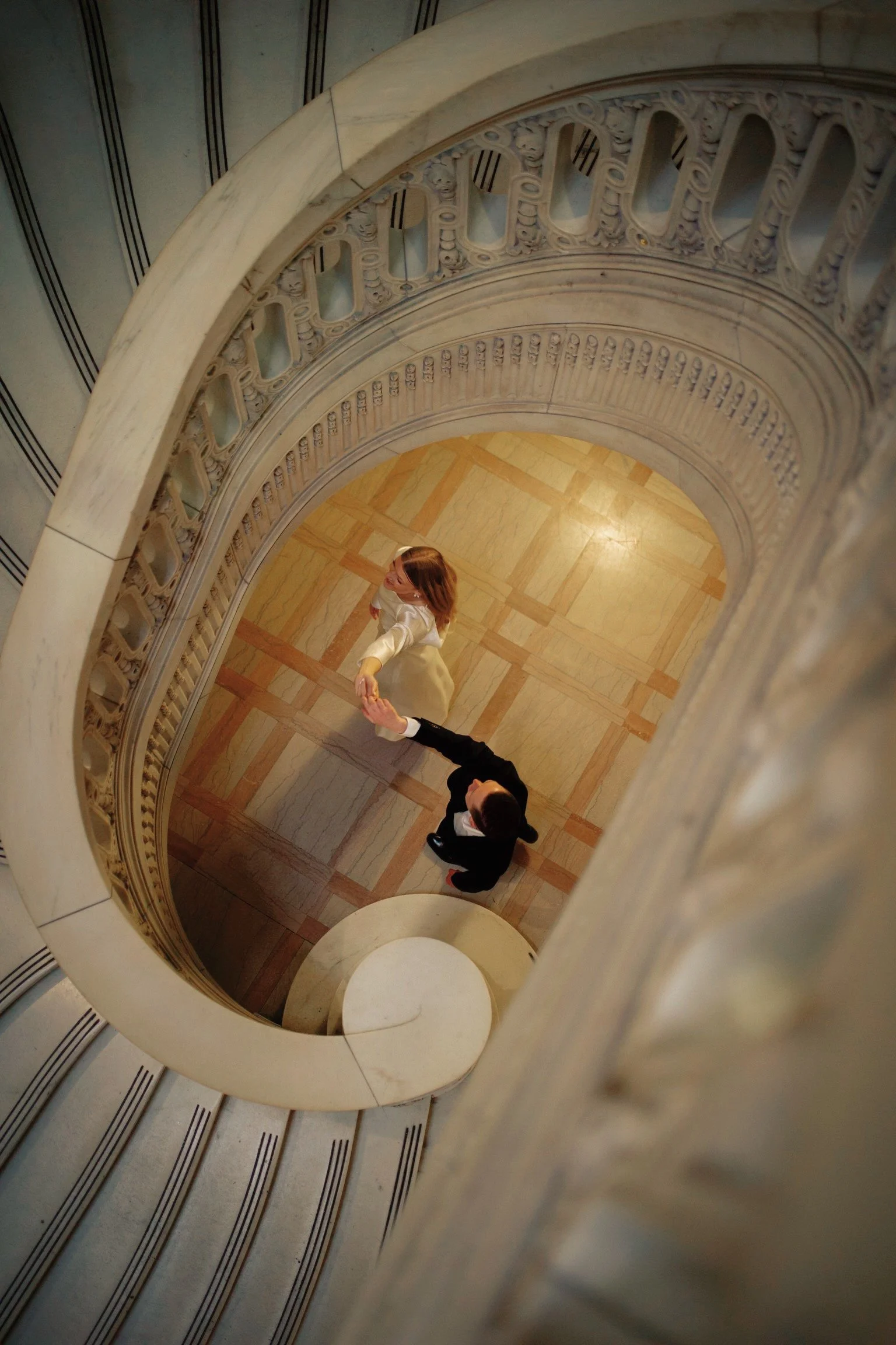 An aerial view of a spiral staircase with ornate railing, showing a man and a woman holding hands on the staircase's floor in a decorative indoor setting.