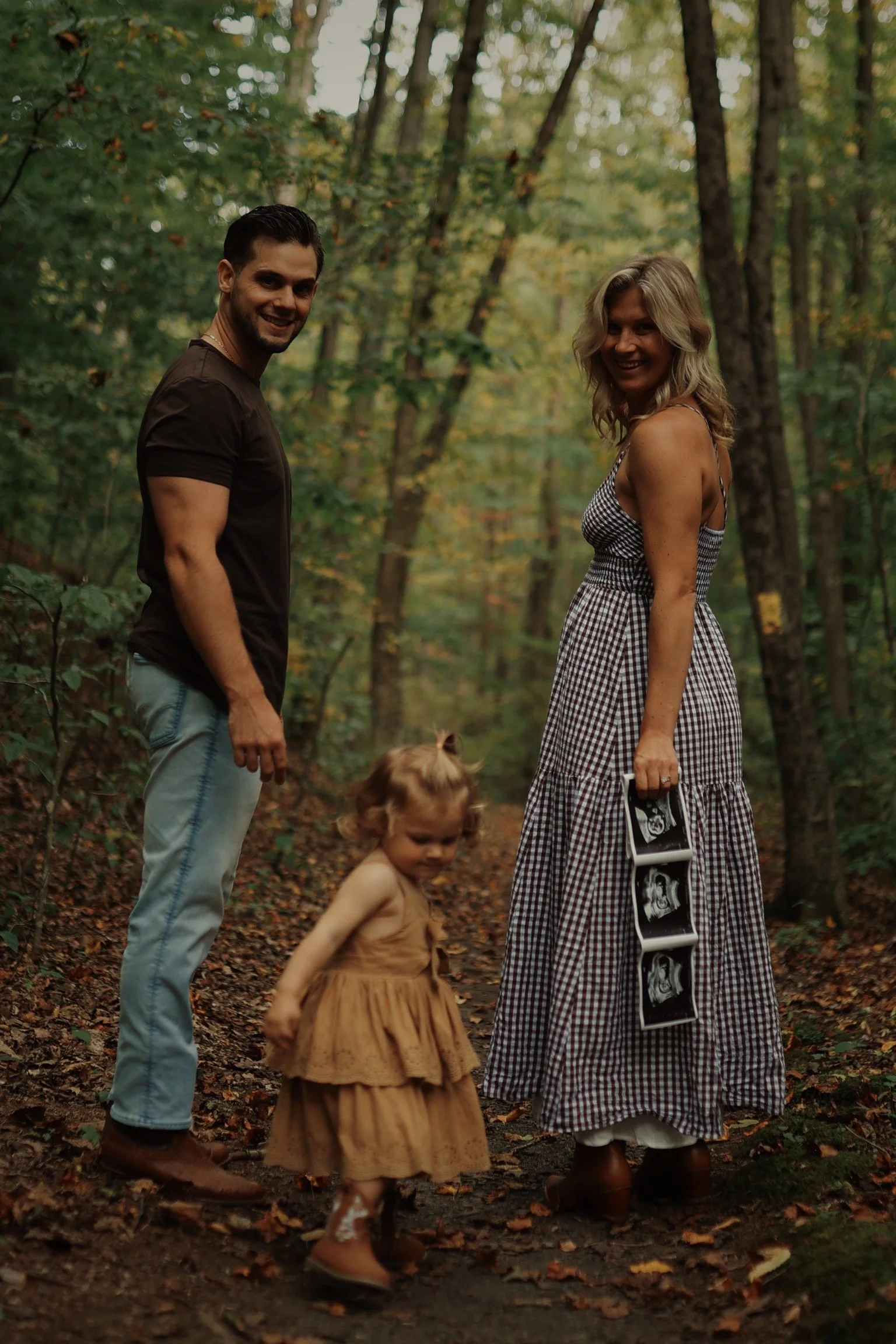 A family of three standing on a forest trail. The woman is holding ultrasound images, and the man and young girl are looking at the camera.
