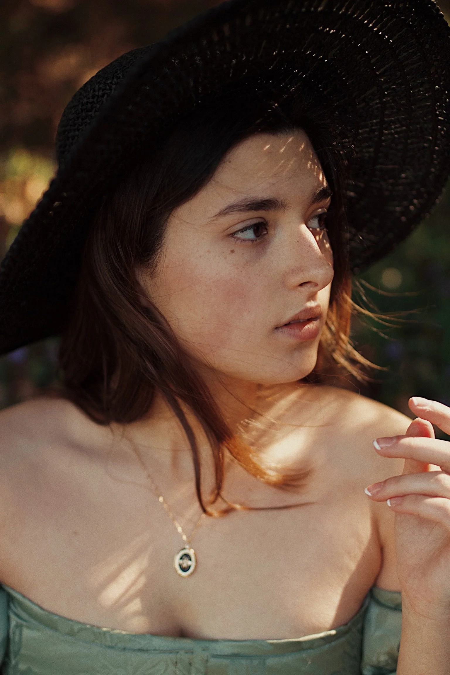 A young woman with dark hair wearing a wide black hat and an off-shoulder top, looking to the side, with sunlight casting patterns on her face and a blurred outdoor background.