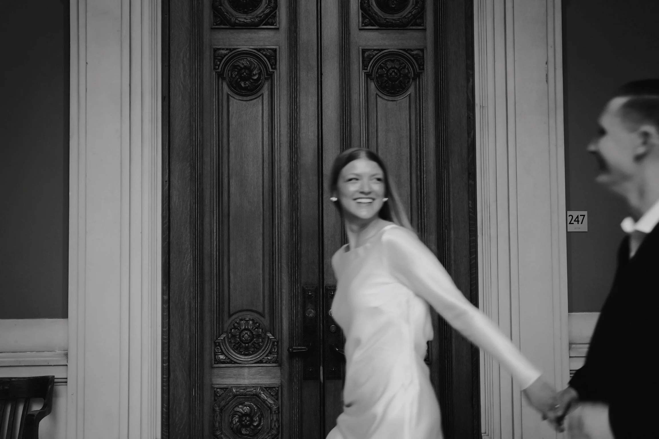 A woman in a white dress and a man in a dark suit holding hands, smiling, in an elegant room with dark wooden doors and ornate trim. The woman is looking back with a smile.
