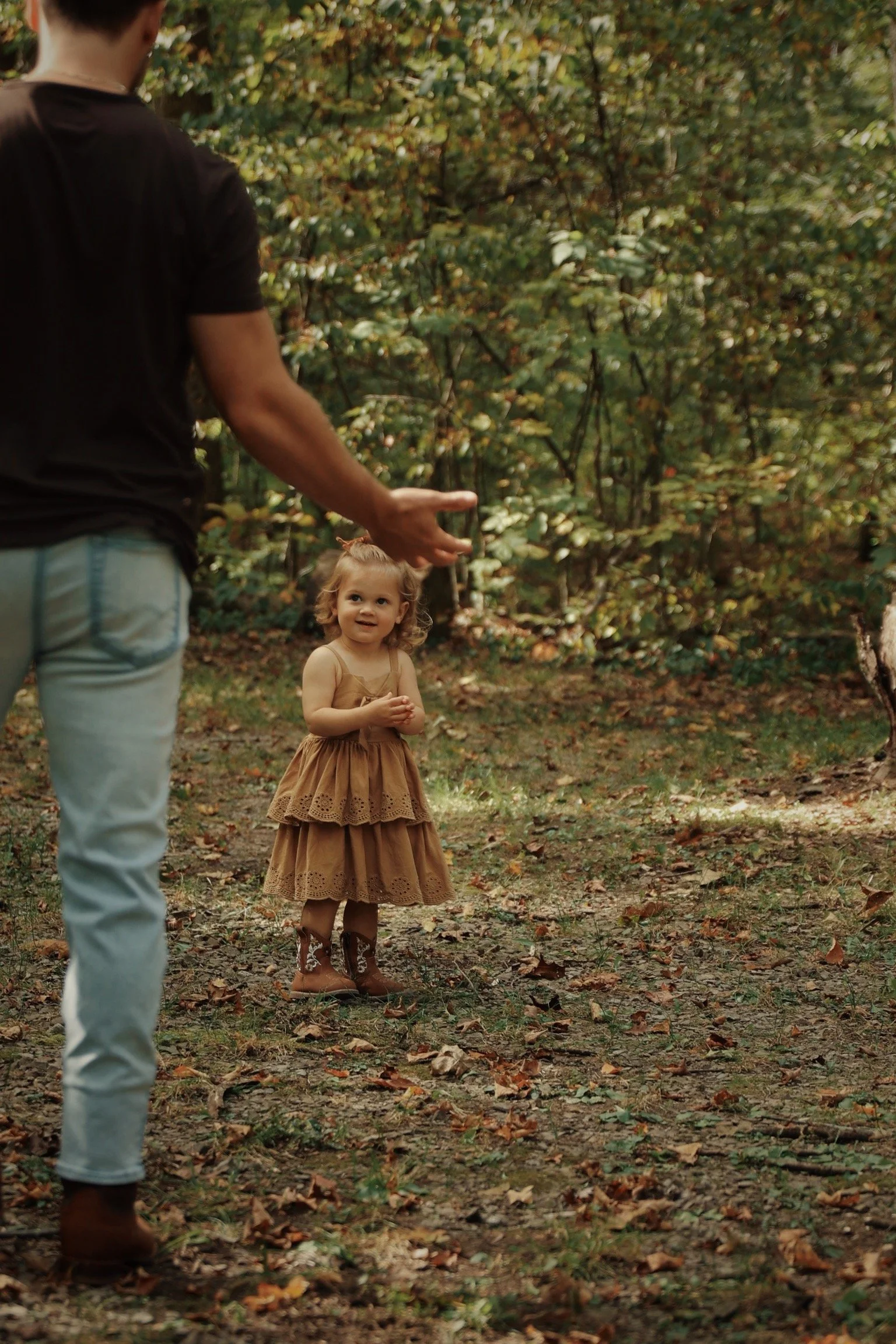 A young girl standing outdoors in a wooded area, wearing a tan dress and cowboy boots, looking up at a man holding a smartphone.