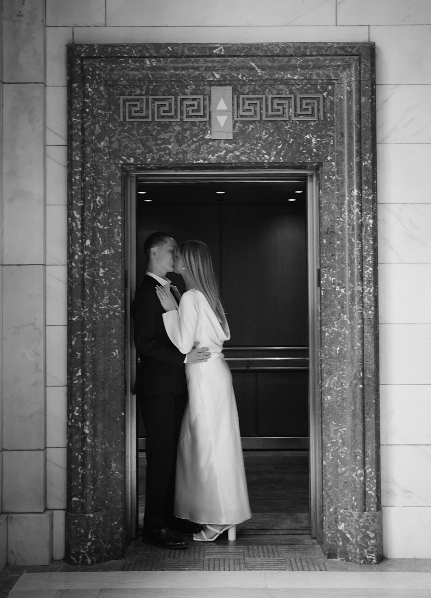 A black-and-white photo of a couple in formal attire sharing an intimate moment in an elevator, with the woman wearing a long dress and the man in a suit, framed by marble features.