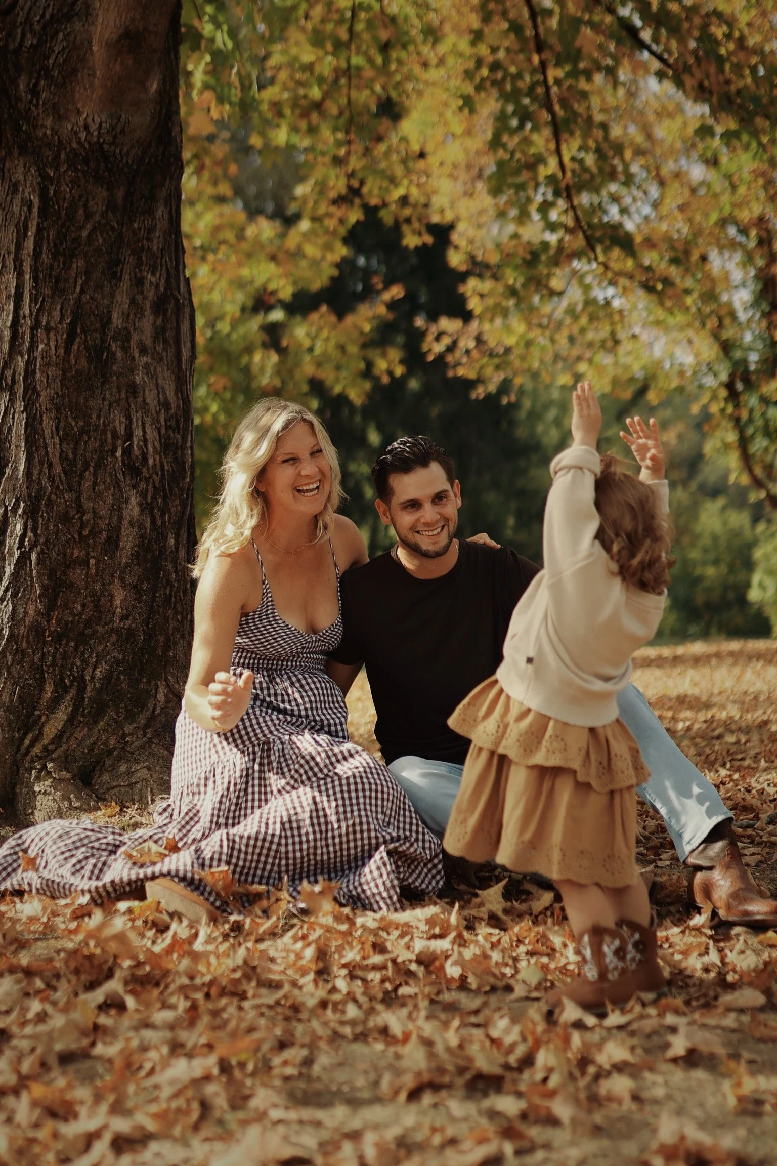A family enjoying autumn outdoors, with mother, father, and young girl playing and laughing on fallen leaves near a large tree with orange and yellow leaves.