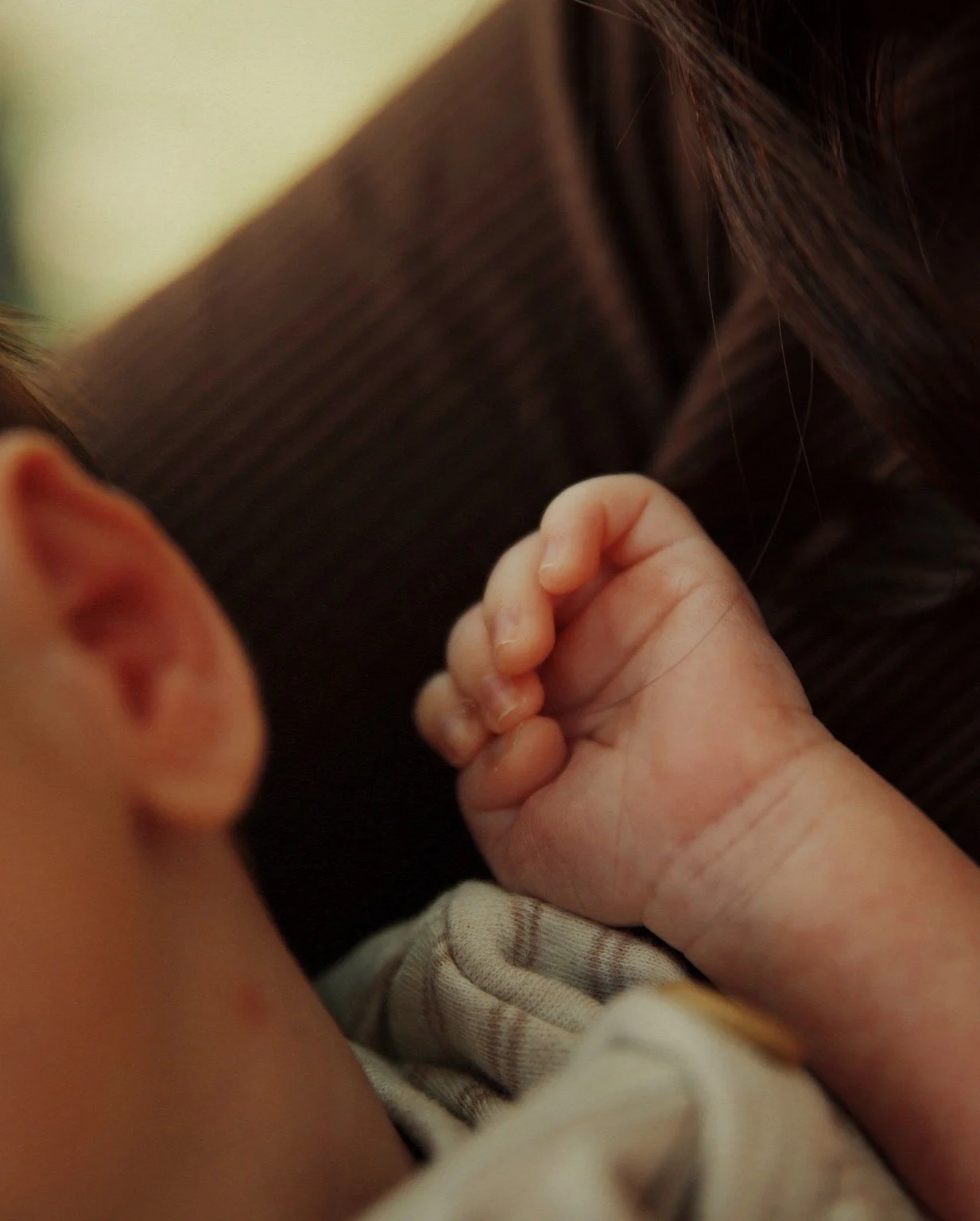 Close-up of a baby's small hand resting on a soft surface, with a blurred background showing part of the baby's face and an adult's arm.