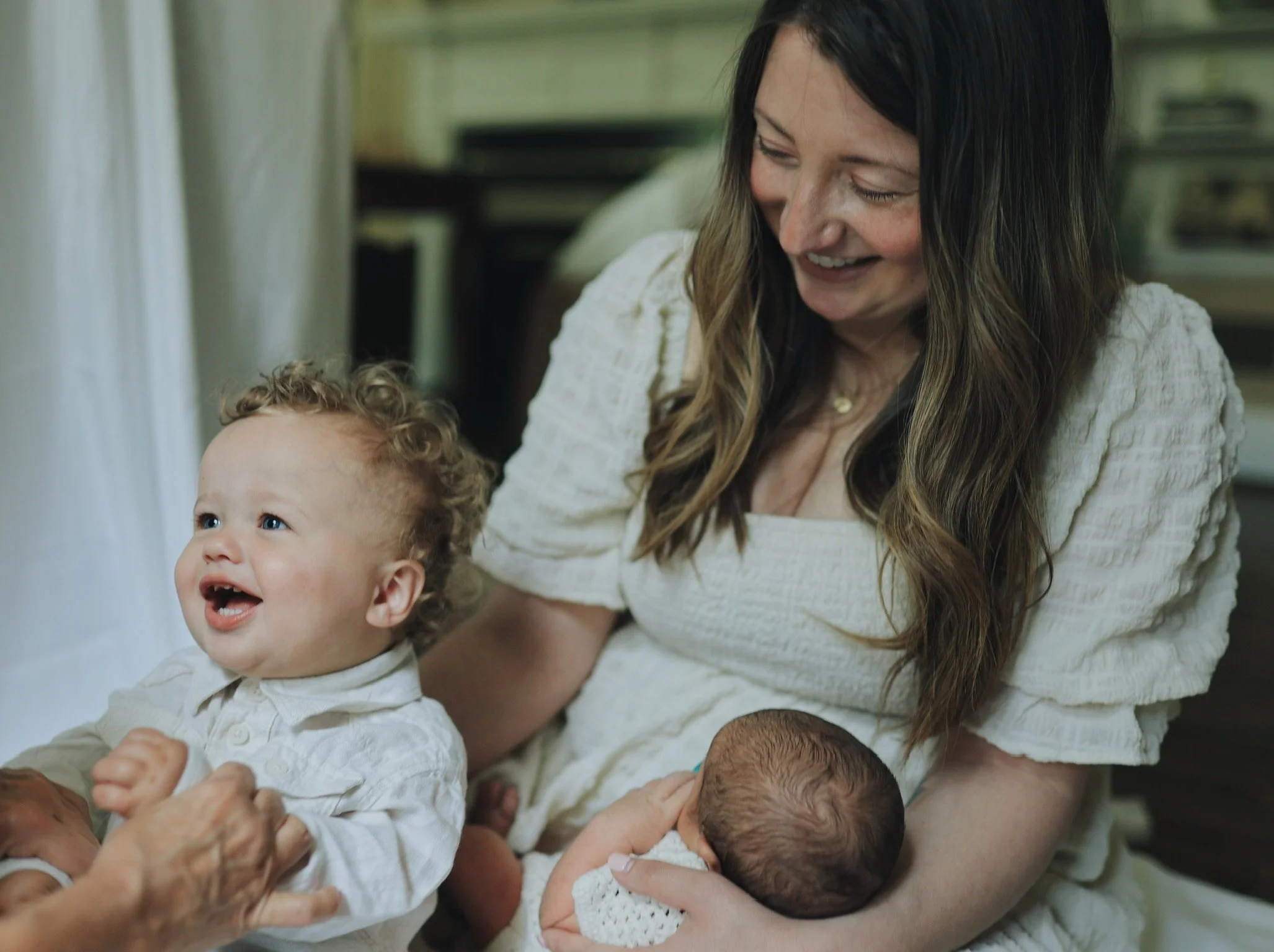 A smiling woman holding a newborn baby in one arm and an older toddler with curly hair in the other, in a warmly lit room.