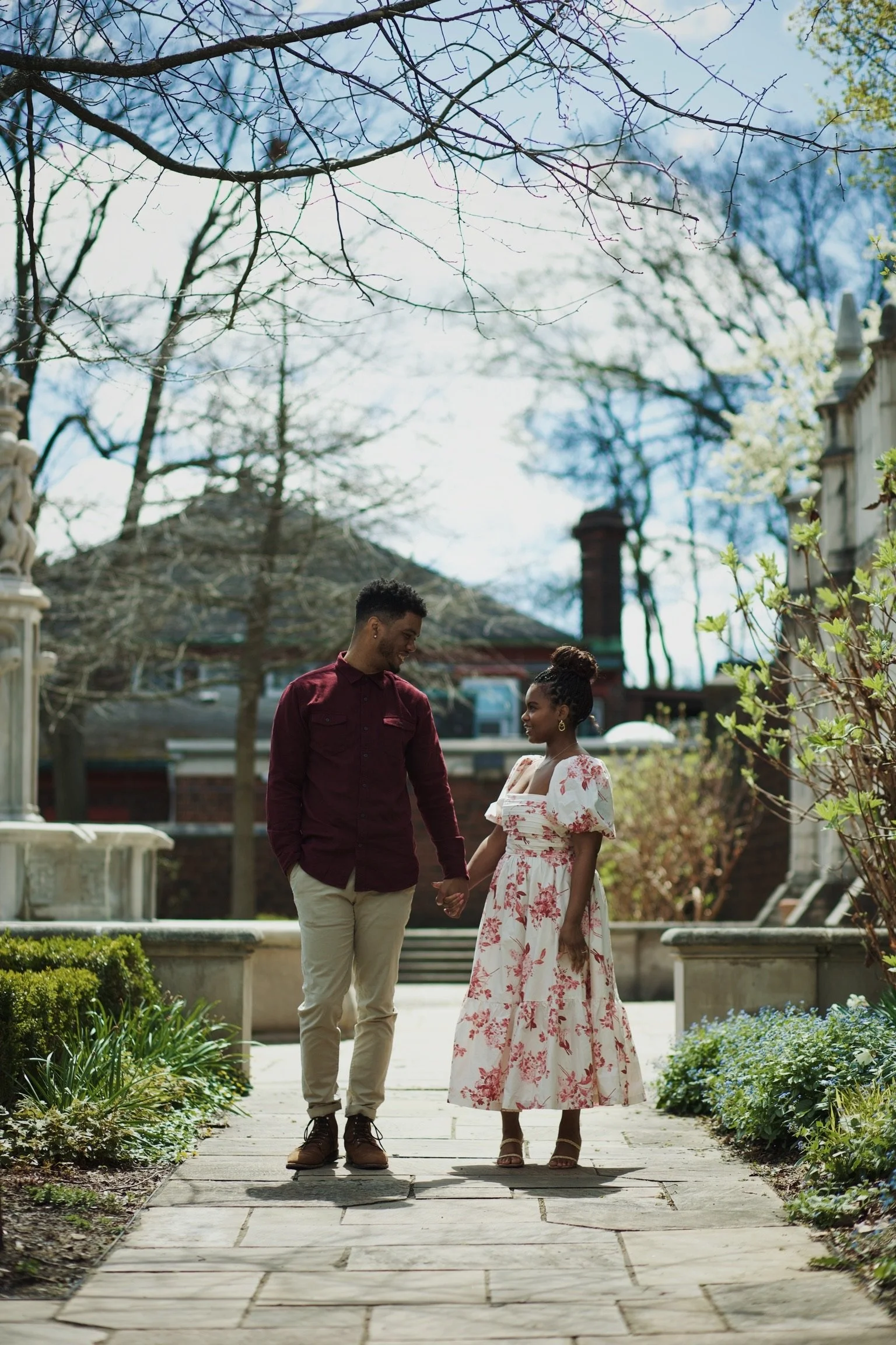 A couple holding hands and walking in a garden on a sunny day, surrounded by trees and plants.