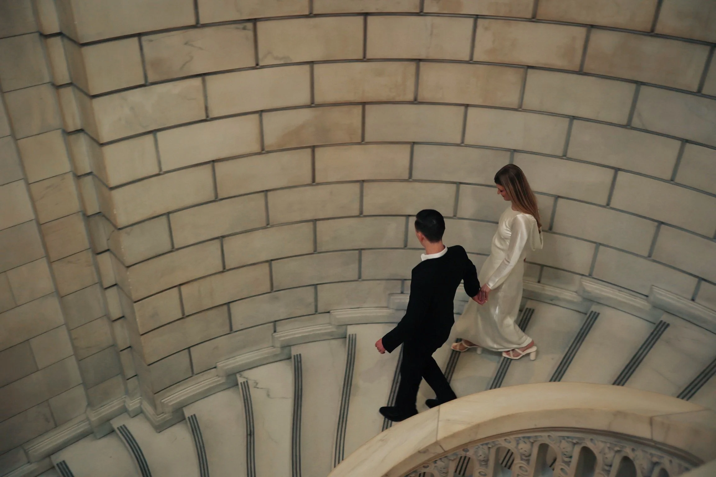 A man and woman holding hands walking down a curved marble staircase in an elegant building with stone walls.