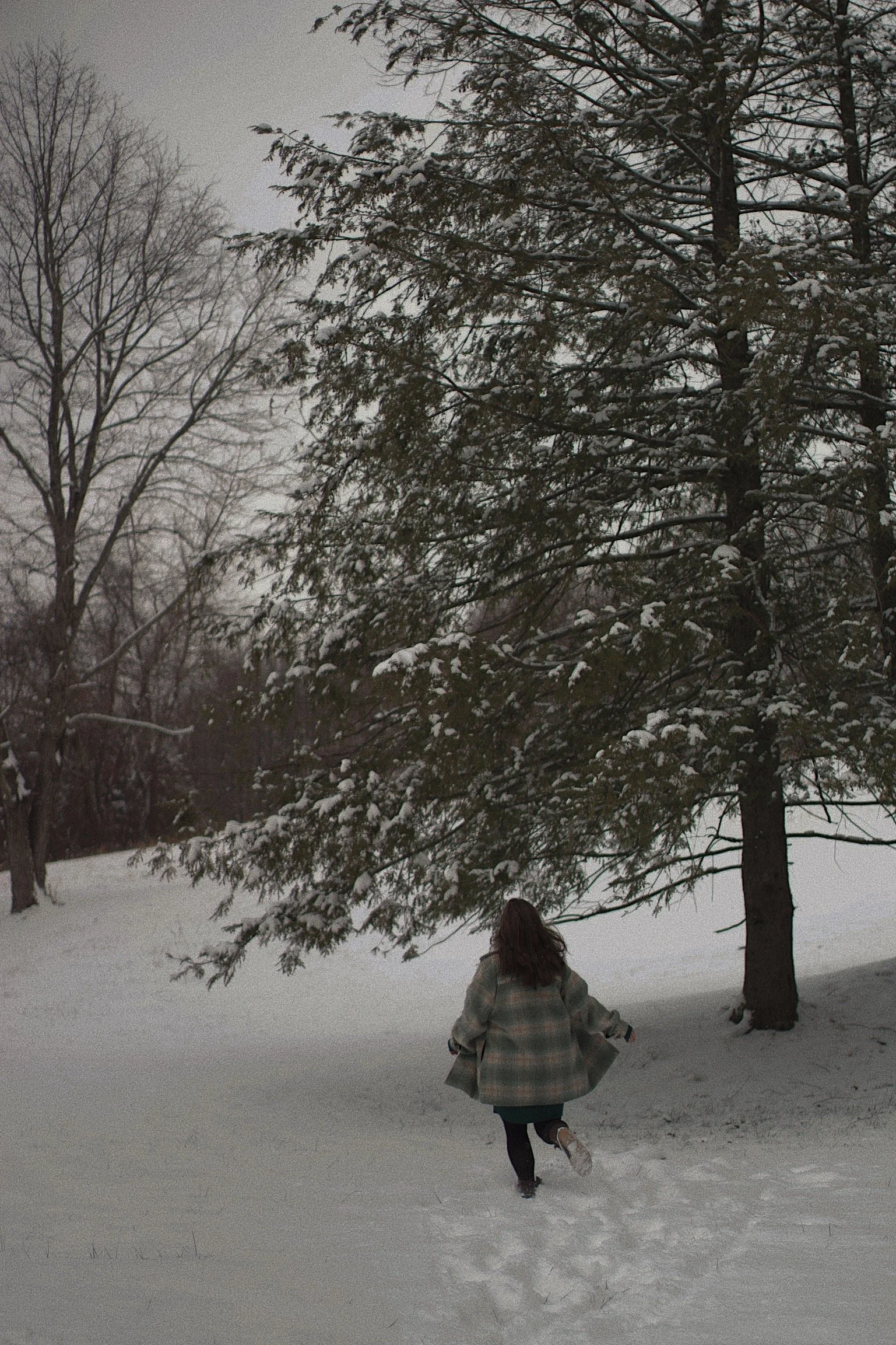 A girl in a plaid coat running through snow near a large snow-covered tree in a winter park.