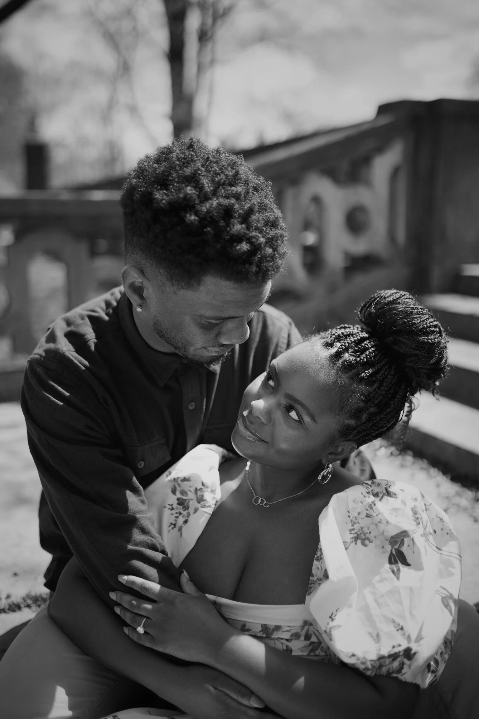 A black and white photo of a man and woman outdoors, gazing into each other's eyes, with the man holding the woman on his lap, and a fence and trees in the background.
