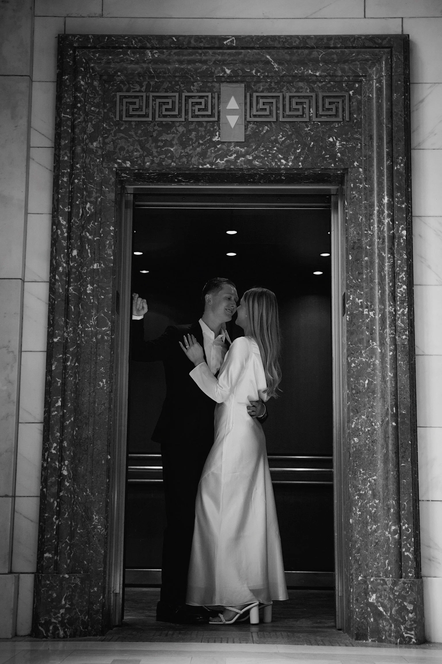 A couple kissing inside an elevator, with the woman wearing a long dress and heels and the man in a suit, framed by marble walls.