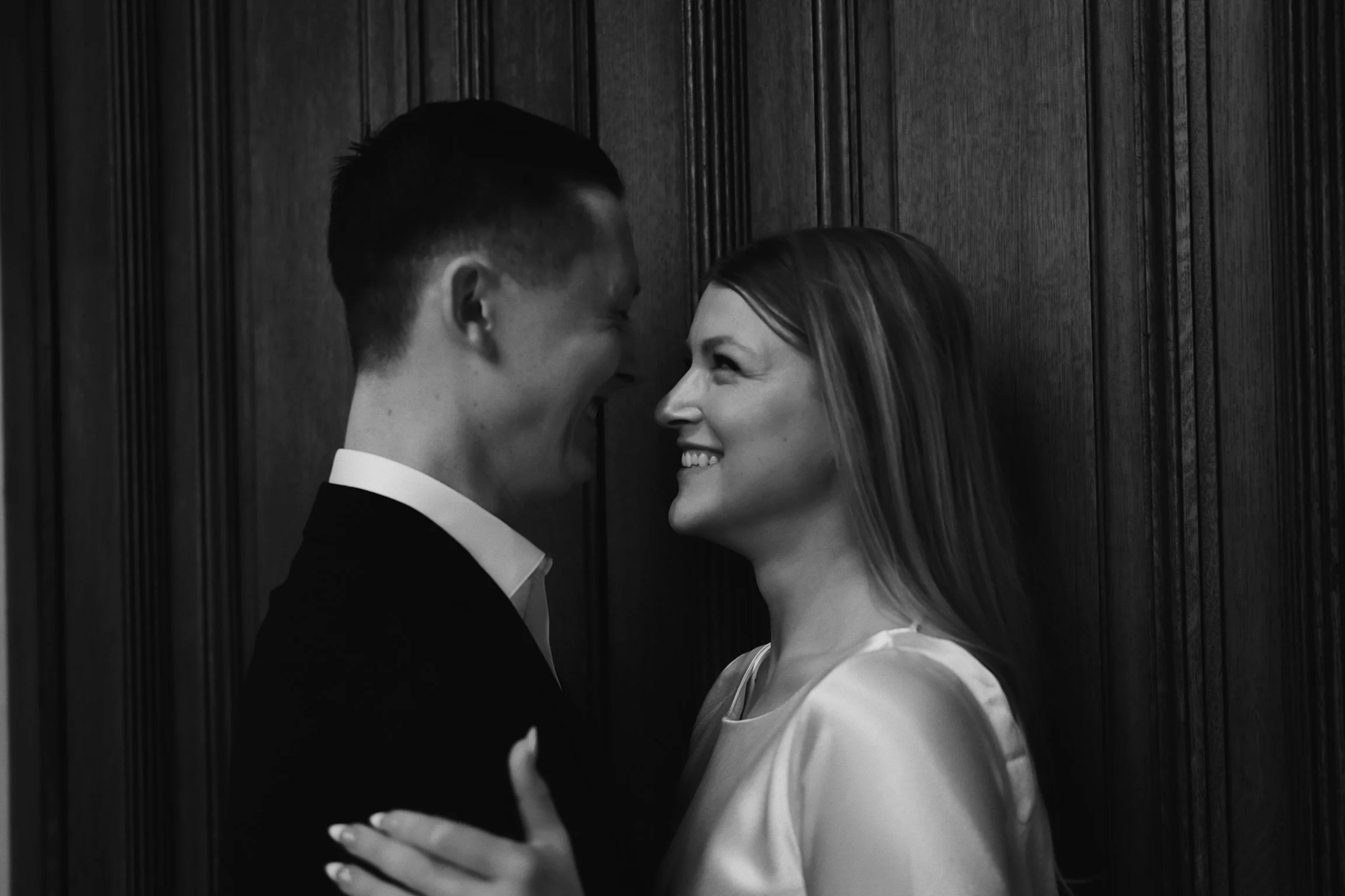 A black and white photo of a smiling couple facing each other closely indoors, with a dark wooden wall in the background.