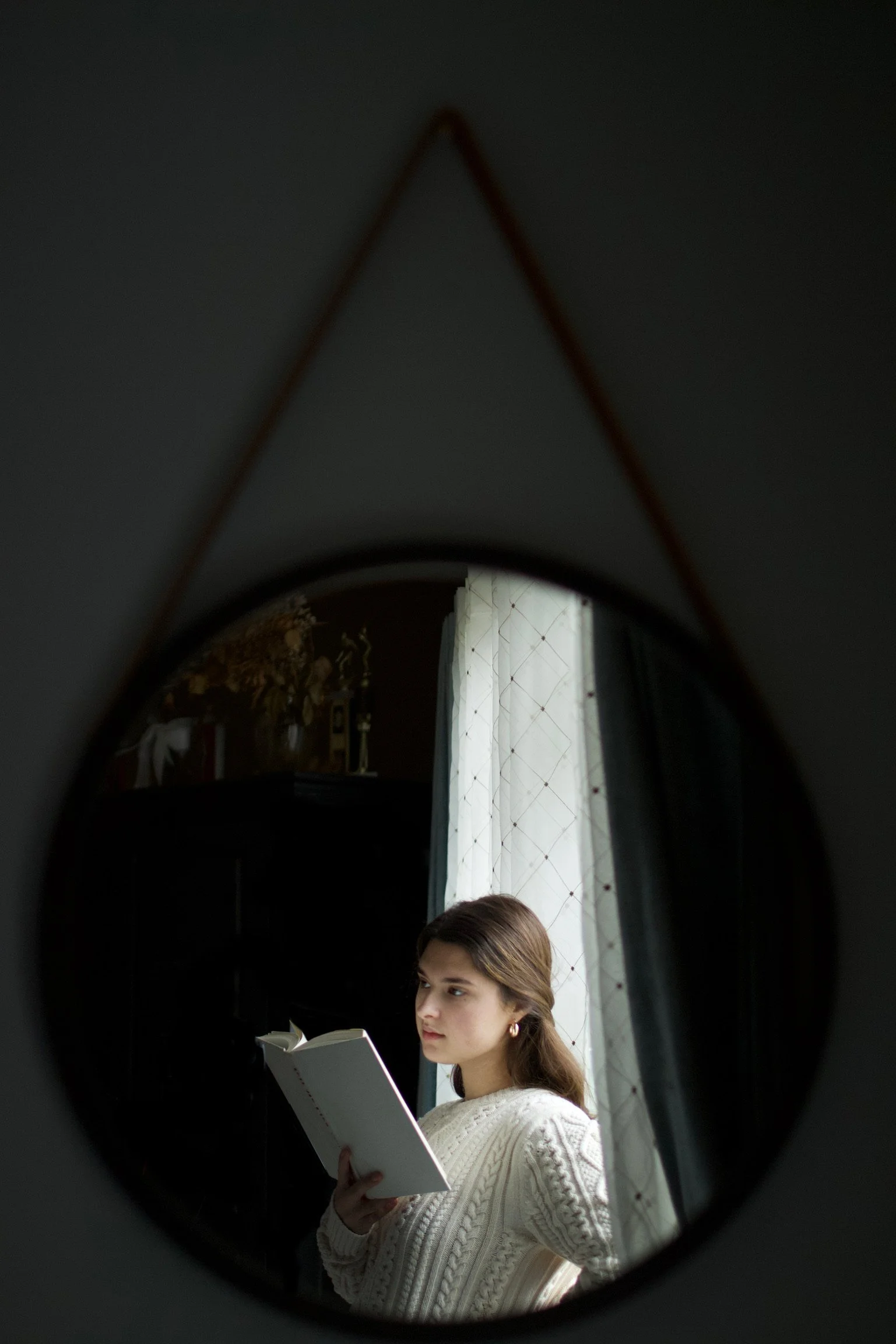 A young woman with brown hair, wearing a white cable-knit sweater and hoop earrings, is seen through a circular mirror, reading a book in a dimly lit room near a window with sheer white curtains.
