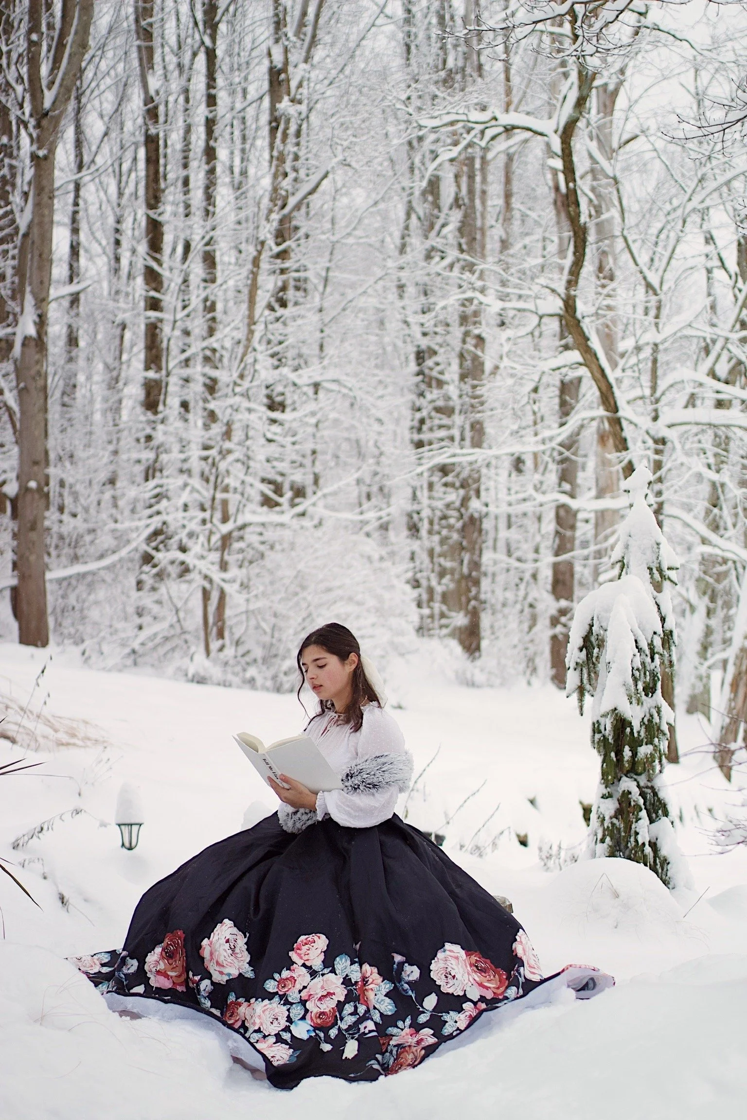 A woman in a long floral skirt and white sweater reading a book while sitting in a snowy forest.