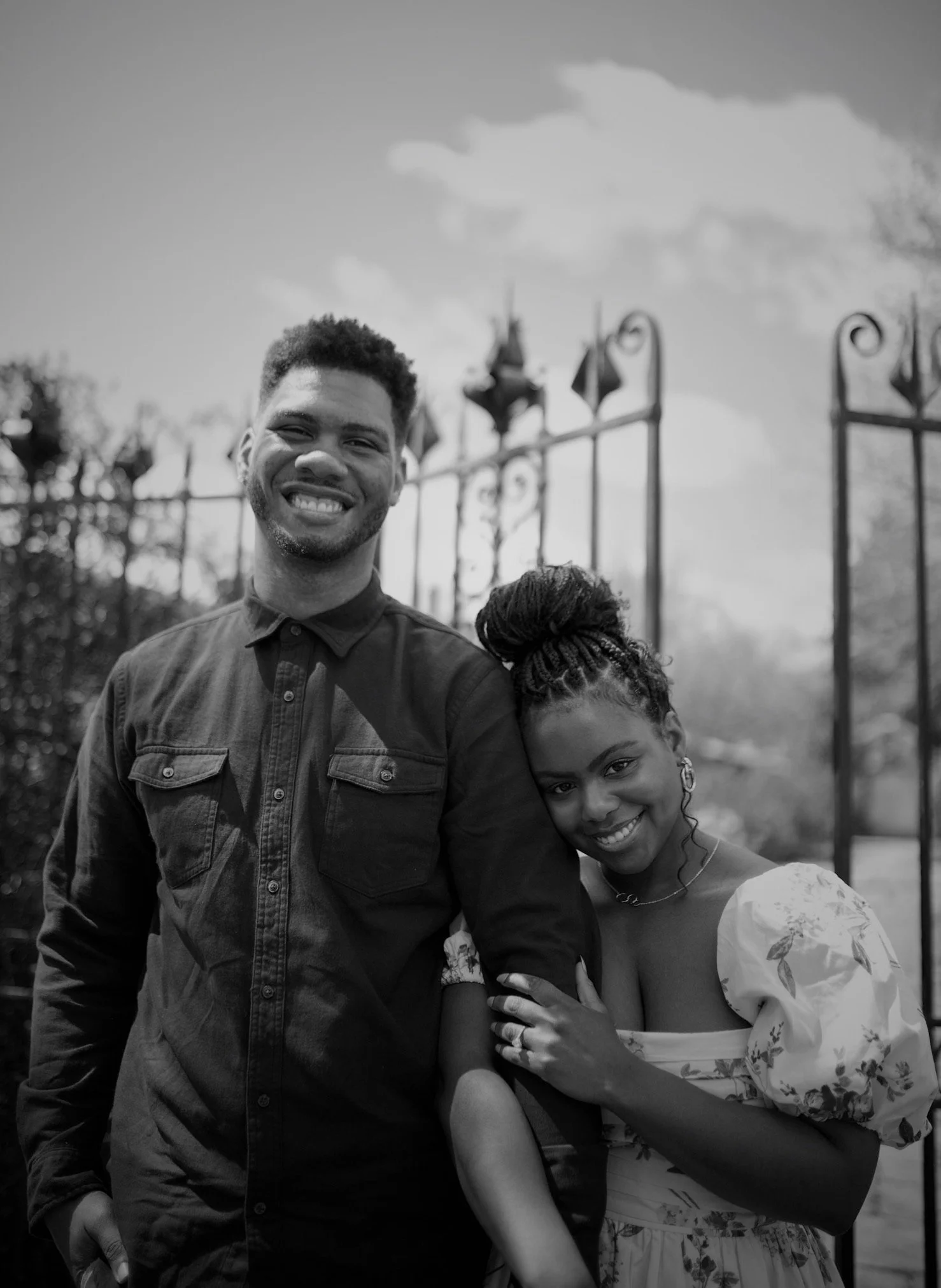 A smiling man and woman stand close together outdoors near a wrought iron gate. The woman leans her head on the man's shoulder, and they are both looking at the camera. The woman has braided hair and wears a floral dress, while the man has short hair