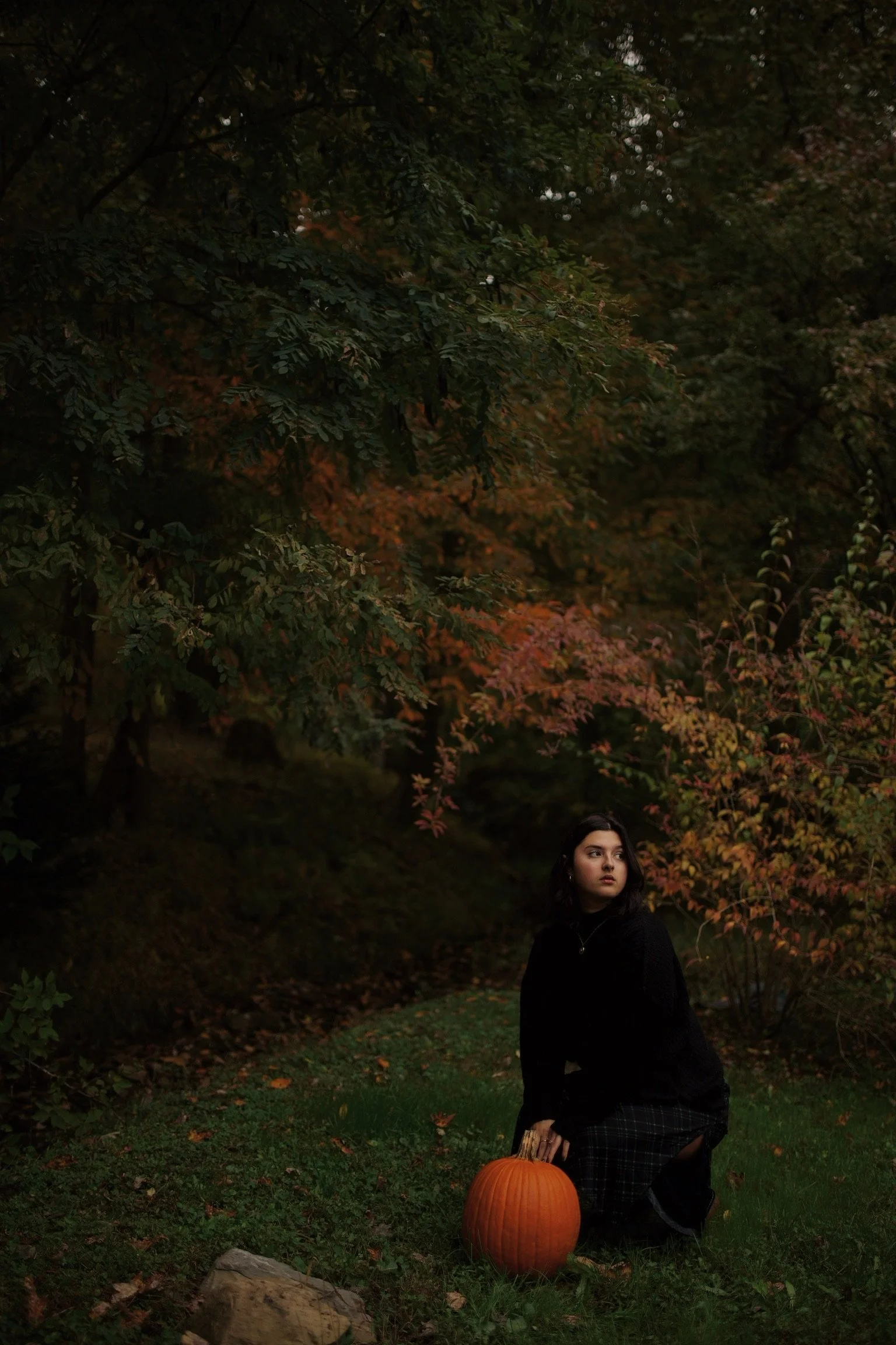 A young woman with dark hair kneeling on grass in a park or wooded area during autumn, holding a large orange pumpkin, surrounded by trees with fall foliage.