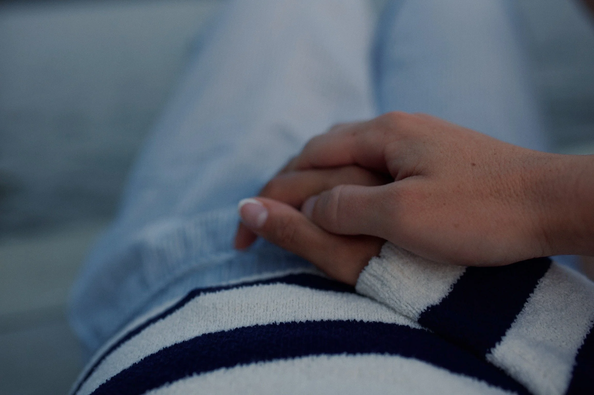 Close-up of two hands clasped together, one hand with a hospital bandage, resting on the leg of a person wearing a striped sleeve.
