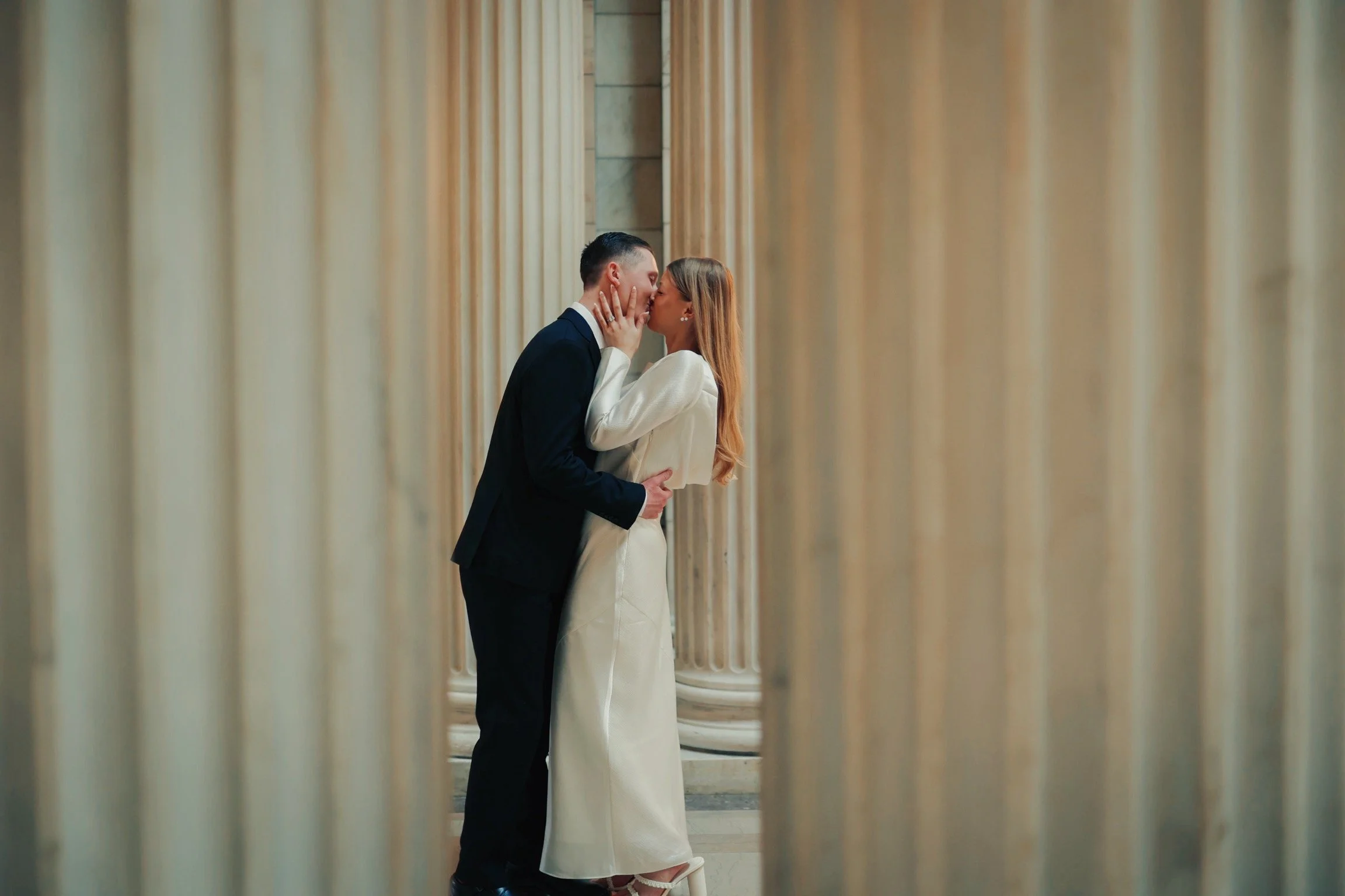 A couple dressed in formal attire sharing an intimate moment between marble columns.