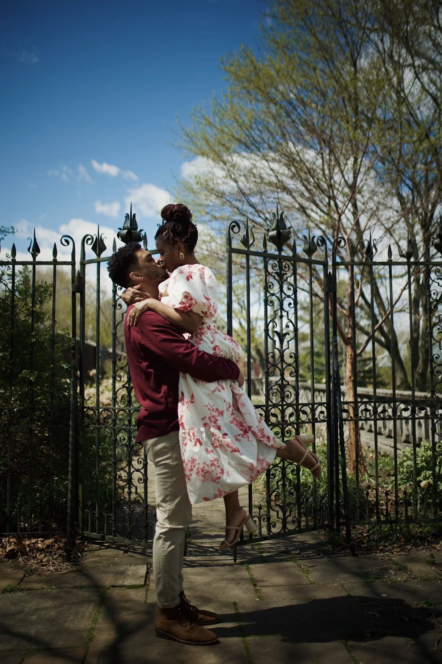 A couple kissing outdoors, with the man lifting the woman. The woman wears a white dress with red floral patterns, and the man wears a maroon shirt and beige pants. There is a black wrought-iron fence and trees in the background under a partly cloudy