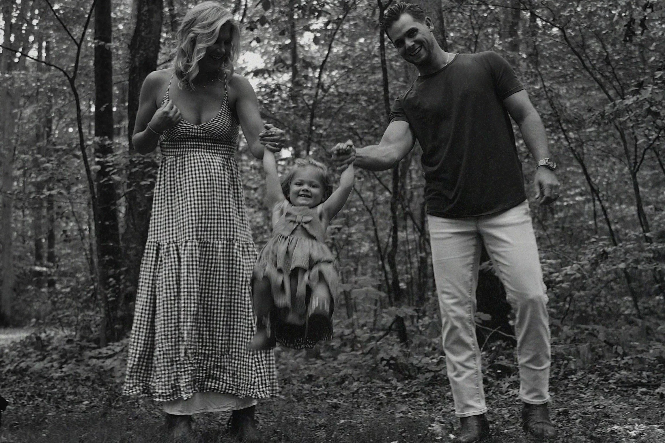A black-and-white photo of a family in a wooded area, with a woman, man, and young girl. The girl is being lifted off the ground by the parents, smiling happily, holding both their hands. The woman is wearing a checkered dress, while the man is in a 