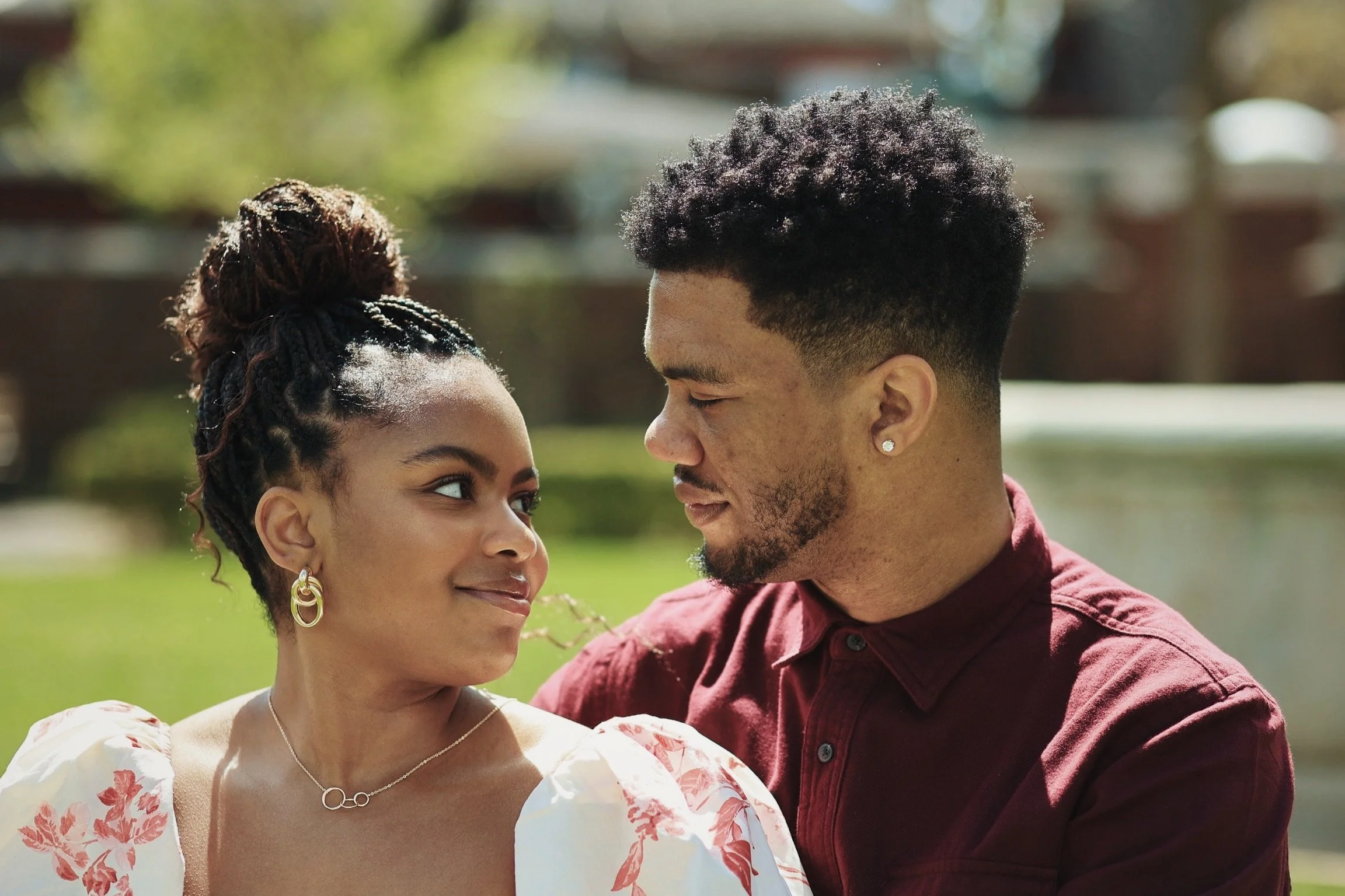 A young woman and a young man look deeply into each other's eyes outdoors. The woman has curly hair in an updo, wearing earrings and a necklace, and a white dress with pink floral patterns. The man has short, curly hair, a beard, and is wearing a mar