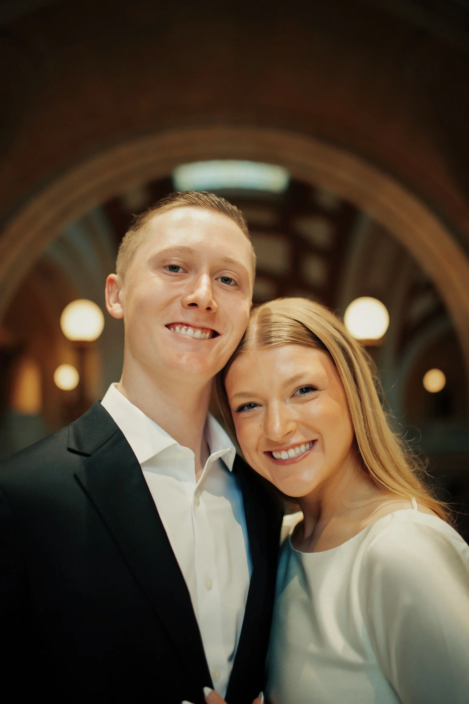 A smiling couple, a man in a black suit and white shirt and a woman in a white dress, standing close together inside a warmly lit historic building.