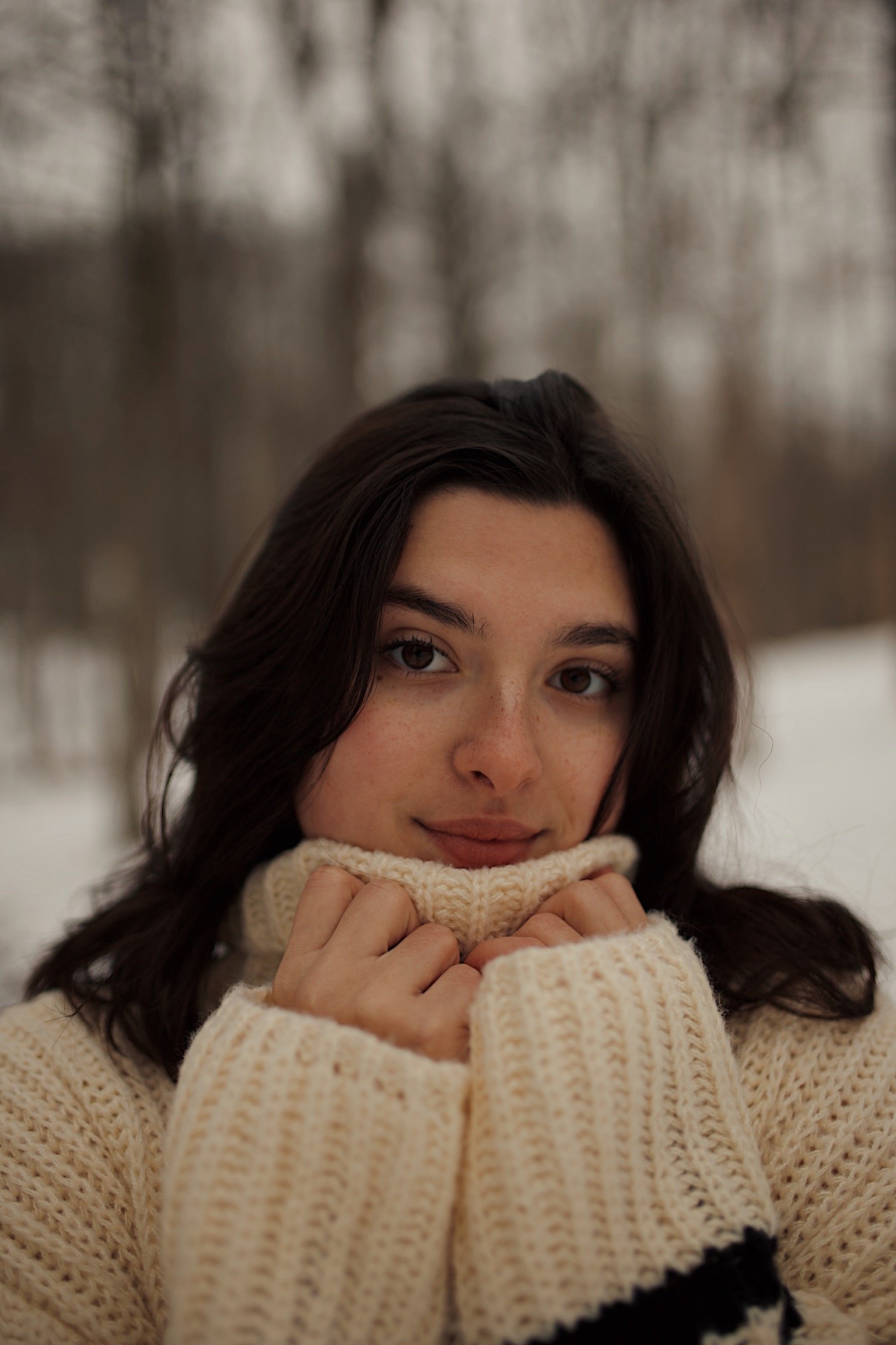 Close-up of a woman with dark hair, wearing a cream-colored knit sweater, pulling the turtleneck up to her chin, outdoors in winter with snow and bare trees in the background.