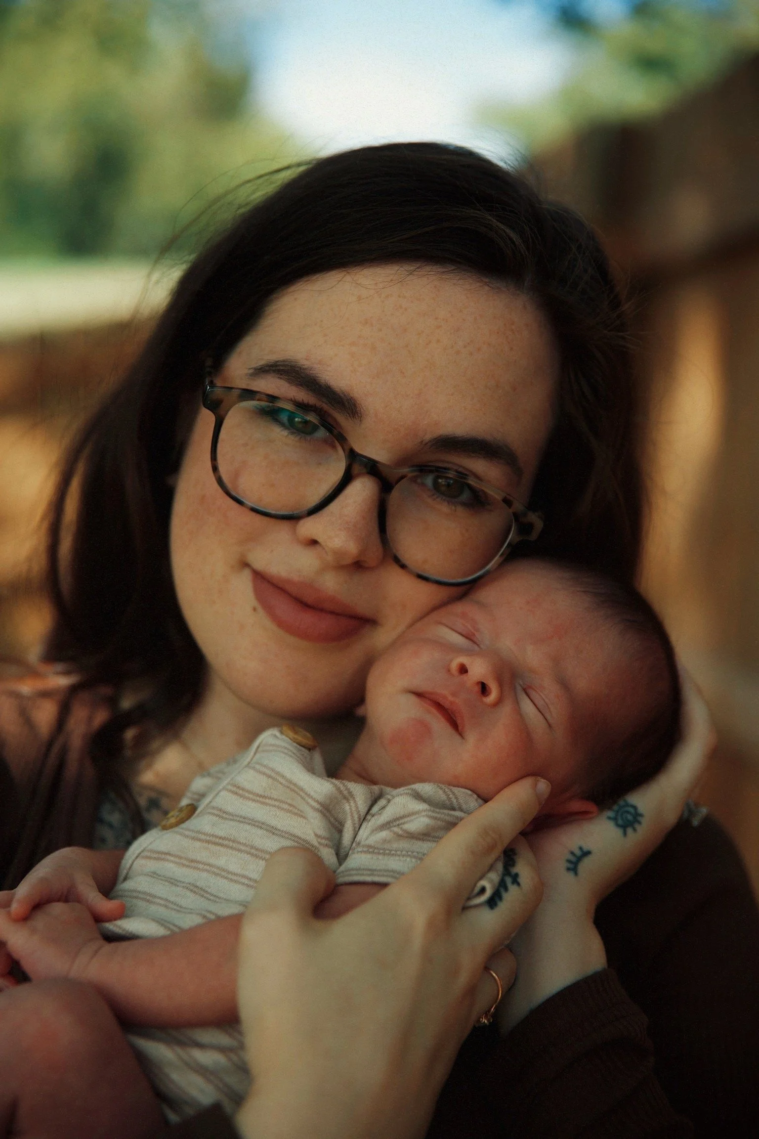 A woman with glasses holding a sleeping newborn baby close to her cheek in an outdoor setting.