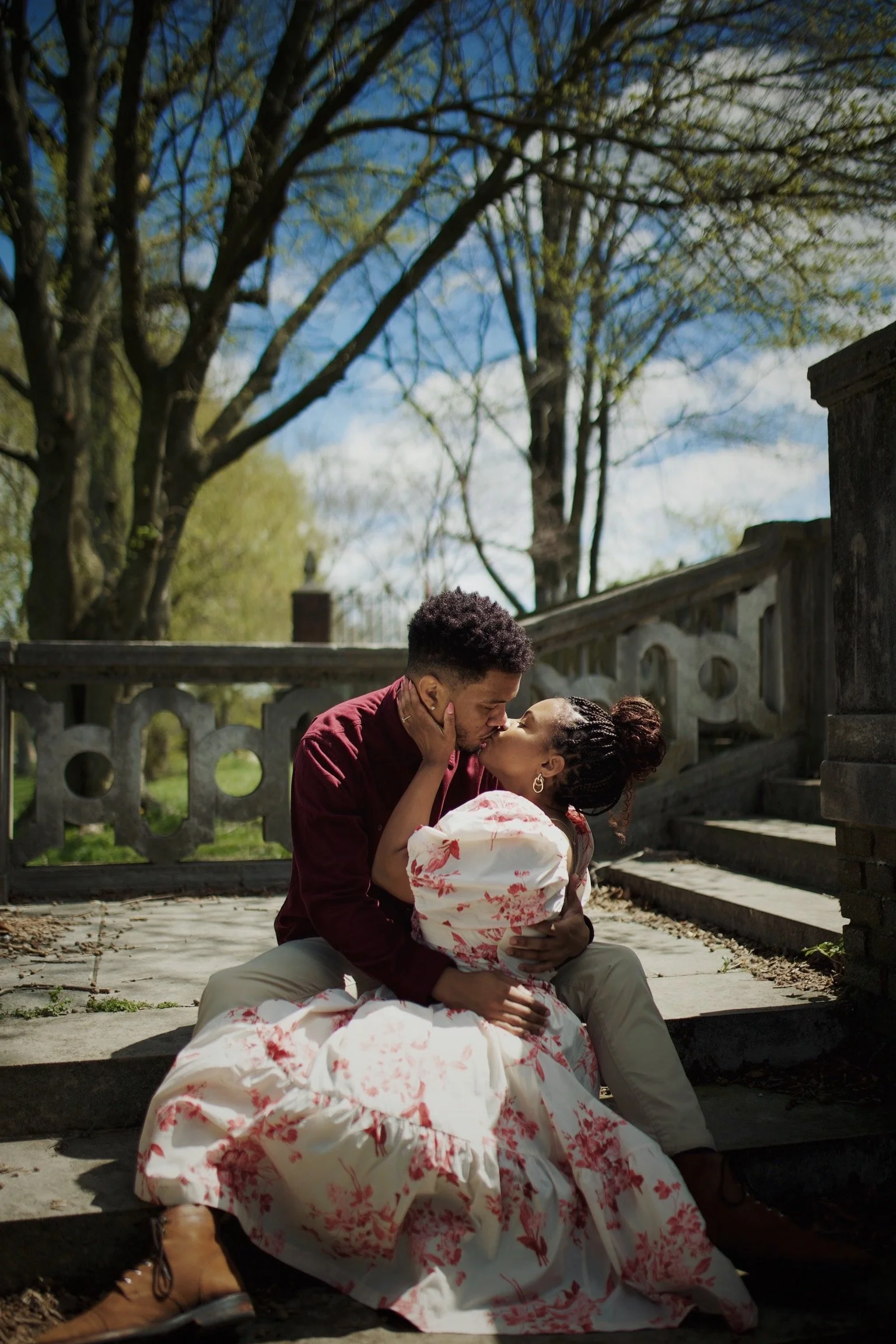 A couple sharing a kiss outdoors, sitting on steps with trees and a blue sky in the background.