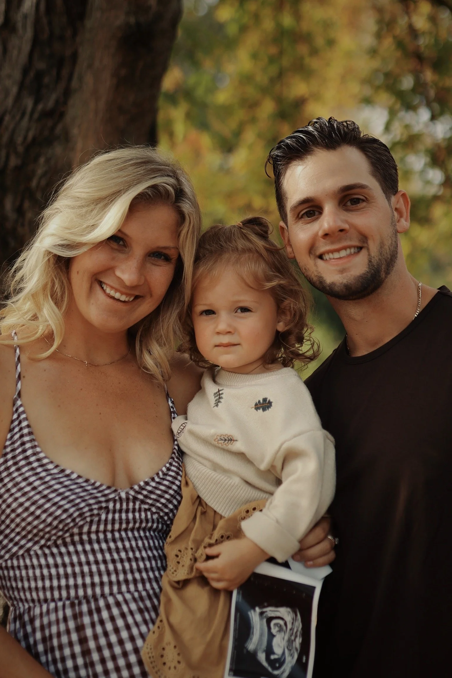 A smiling family of three outdoors near a tree, with the woman on the left, the woman holding a child, and the man on the right, all looking at the camera.