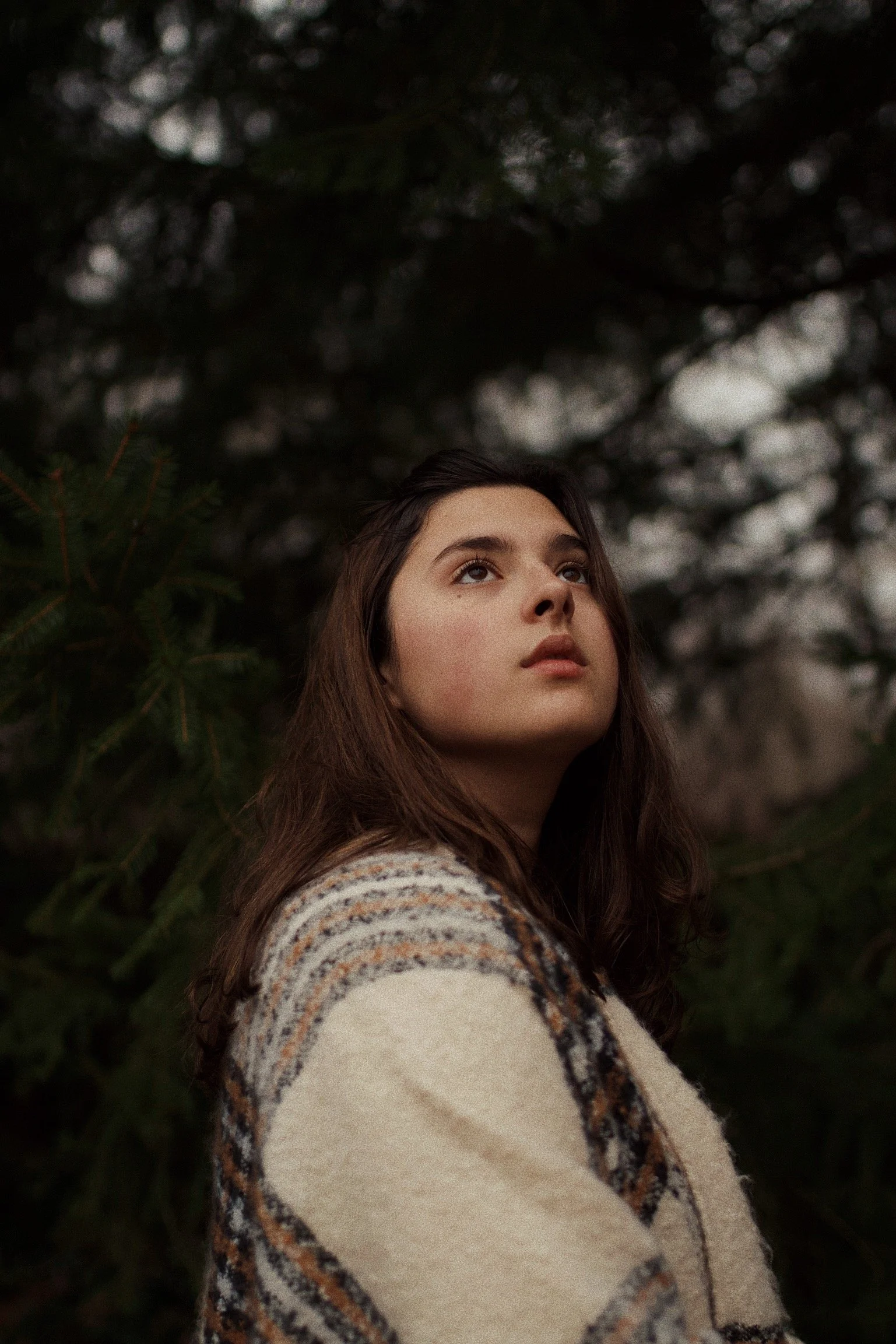 A young woman with long brown hair and fair skin looking upward, standing outdoors among dark green trees.