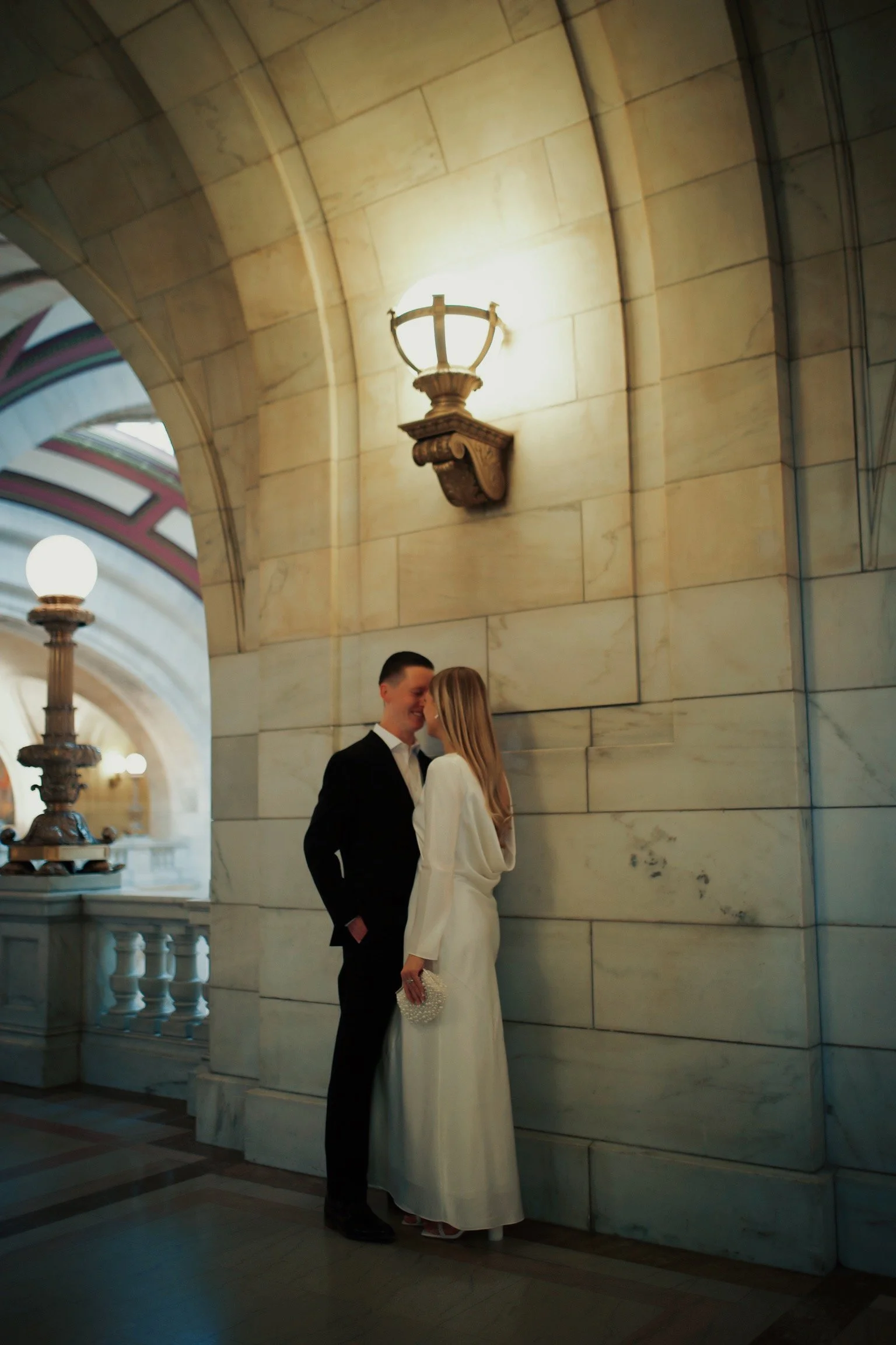 A couple in wedding attire sharing a moment in a grand, marble indoor setting with arched architecture and wall-mounted lamps.
