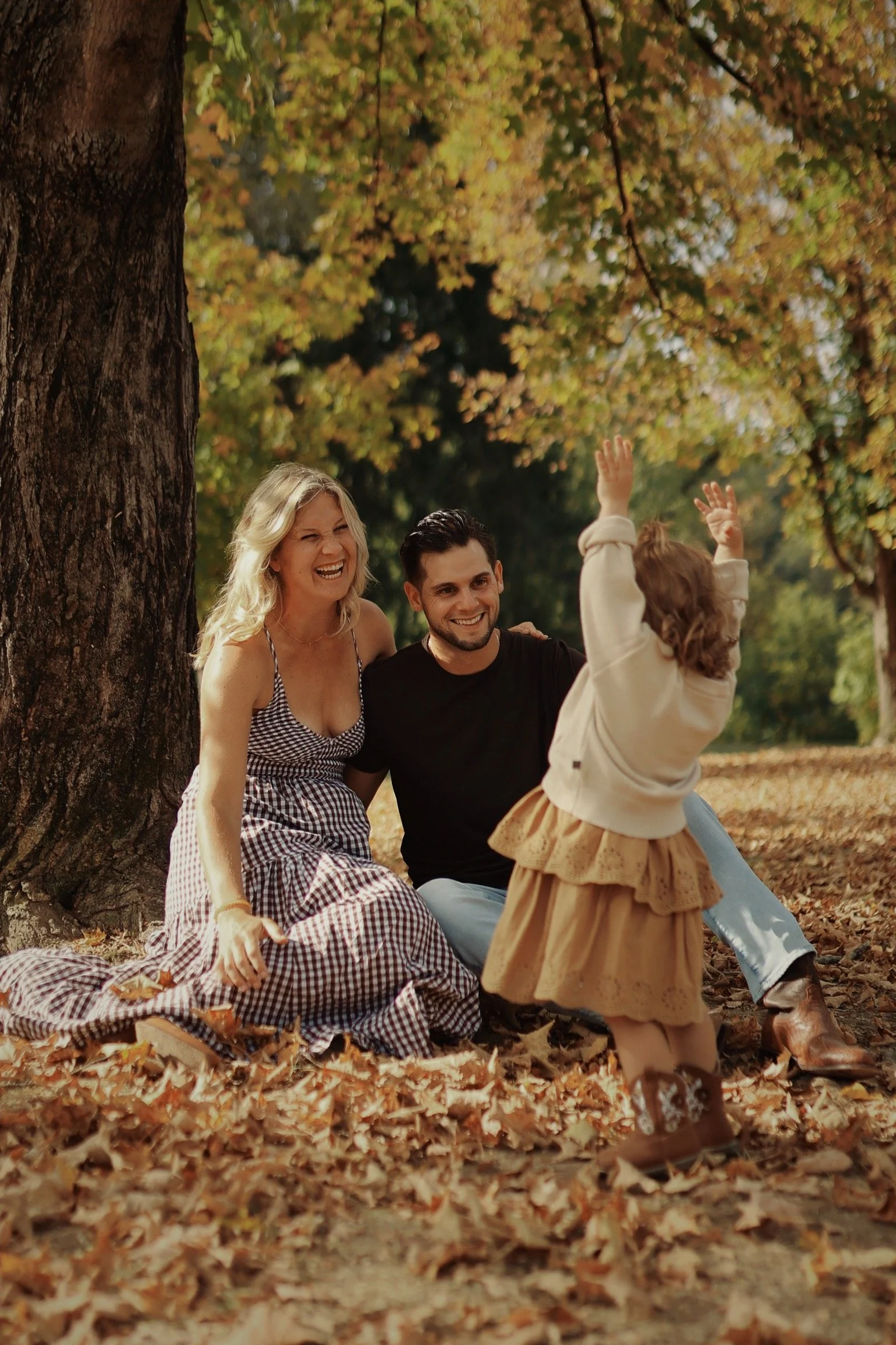 Two adults, a woman in a checkered dress and a man in a t-shirt, sitting on the ground under a large tree, smiling and interacting with a young girl in a beige dress and cardigan, raising her arms in the air during autumn time with fallen leaves arou